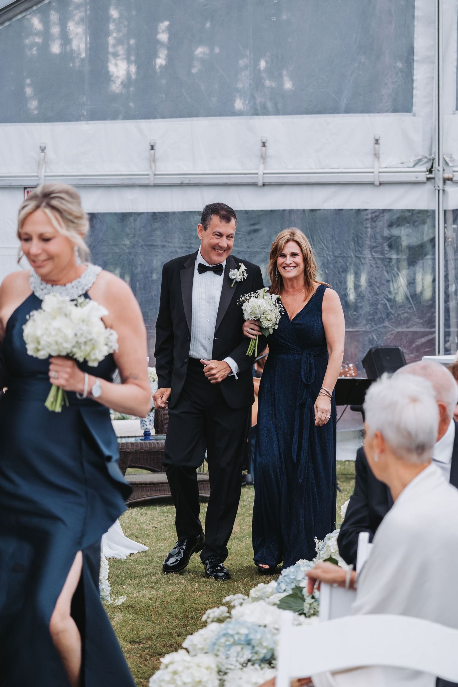 Groomsman in tuxedo escorts bridesmaid in navy gown down outdoor wedding aisle under clear tent.