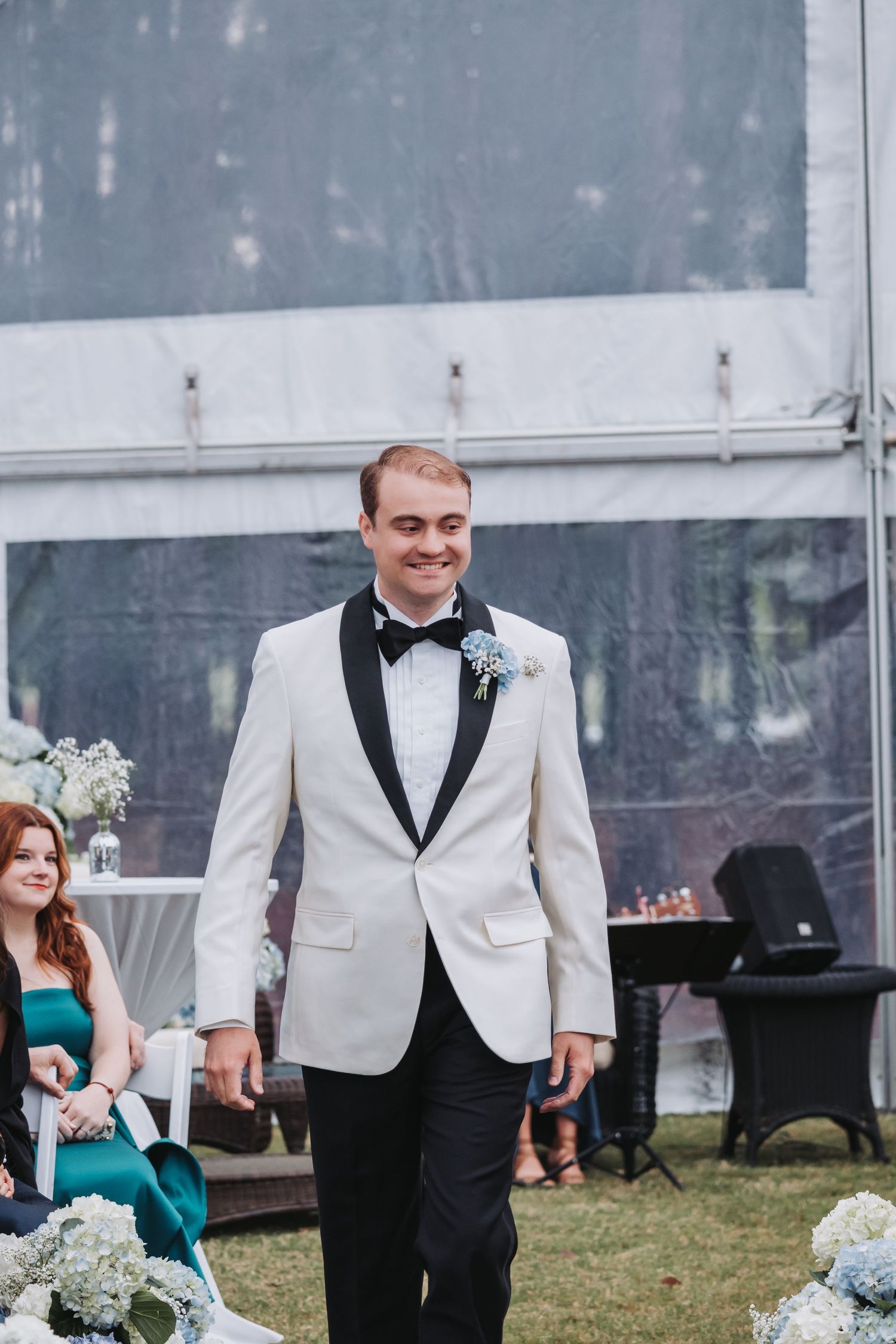 Smiling groom in white tuxedo jacket with black lapels and blue hydrangea boutonniere walks down outdoor wedding aisle.