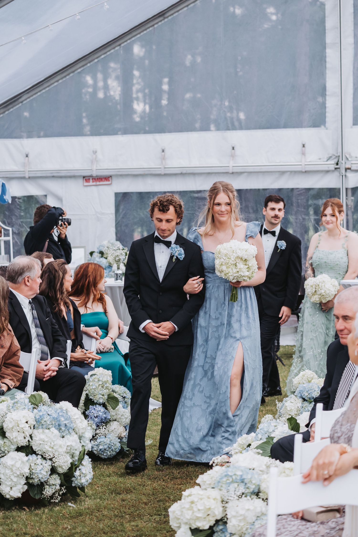 Bridesmaid in blue gown walks down floral aisle lined with white hydrangeas under a clear wedding tent.