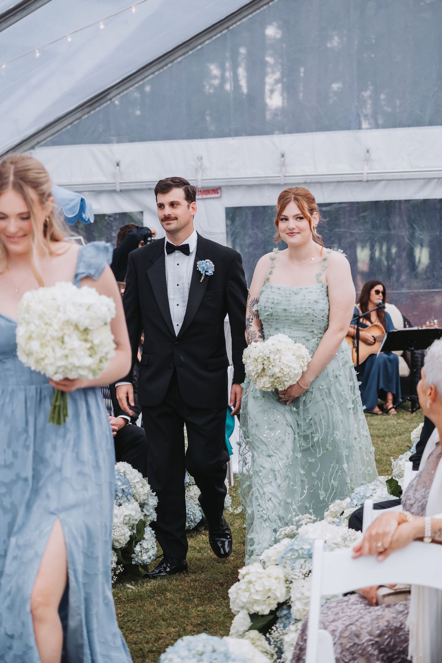 Groomsman in black tuxedo escorts bridesmaid in sage green gown down aisle at outdoor tented wedding.