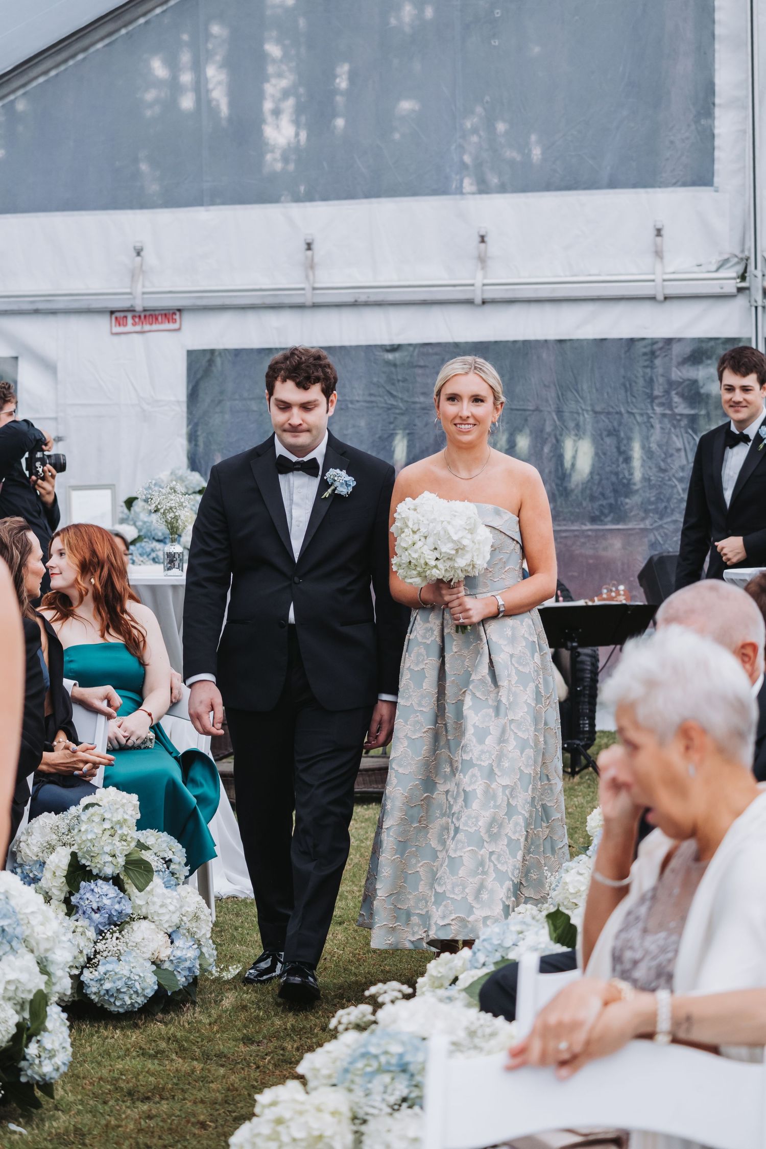 Bride and groomsman walk down outdoor wedding aisle lined with blue and white hydrangeas under a white tent.