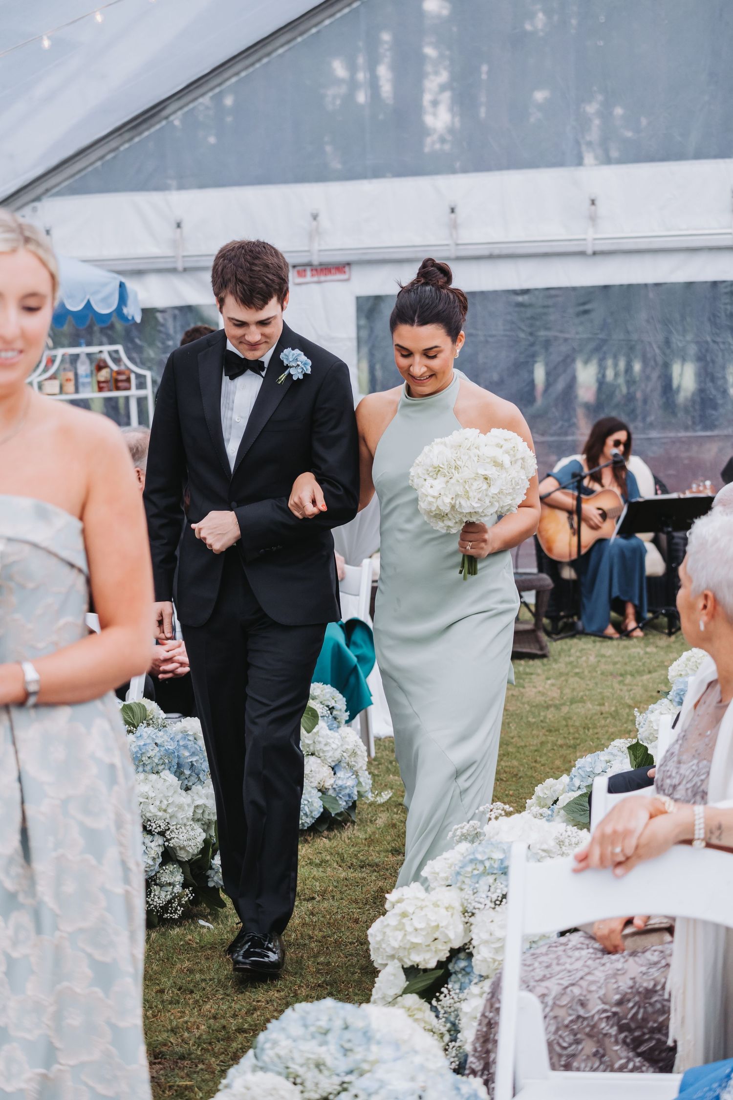 Groomsman escorts bridesmaid in sage green dress down aisle lined with blue hydrangeas at tent ceremony.