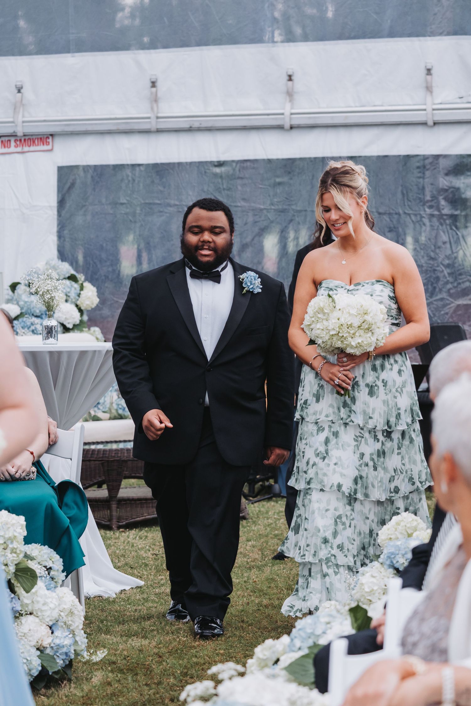 Groomsman in black tuxedo escorts smiling bridesmaid in floral dress down hydrangea-lined wedding aisle.