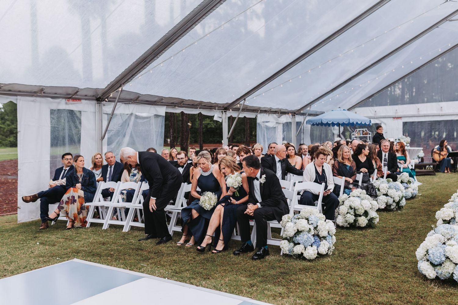 Wedding guests seated in white chairs under clear tent with white hydrangea floral arrangements lining the aisle.