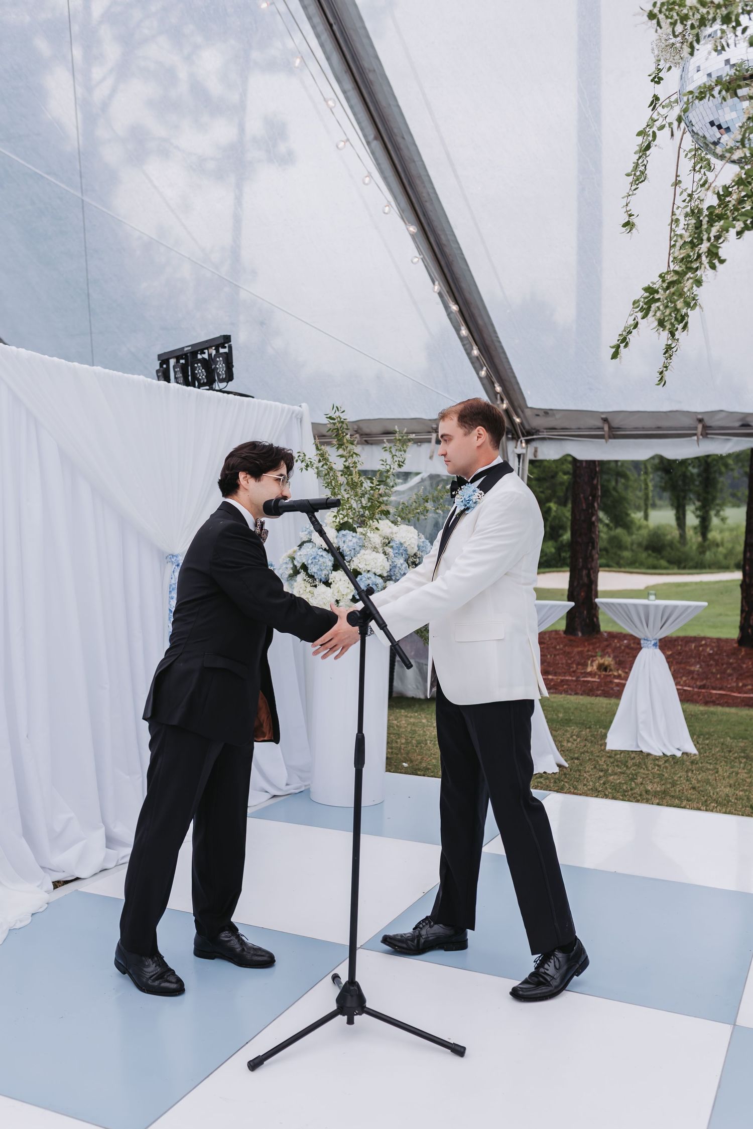 Two men in formal attire arrange floral centerpiece on microphone stand at elegant outdoor wedding ceremony.