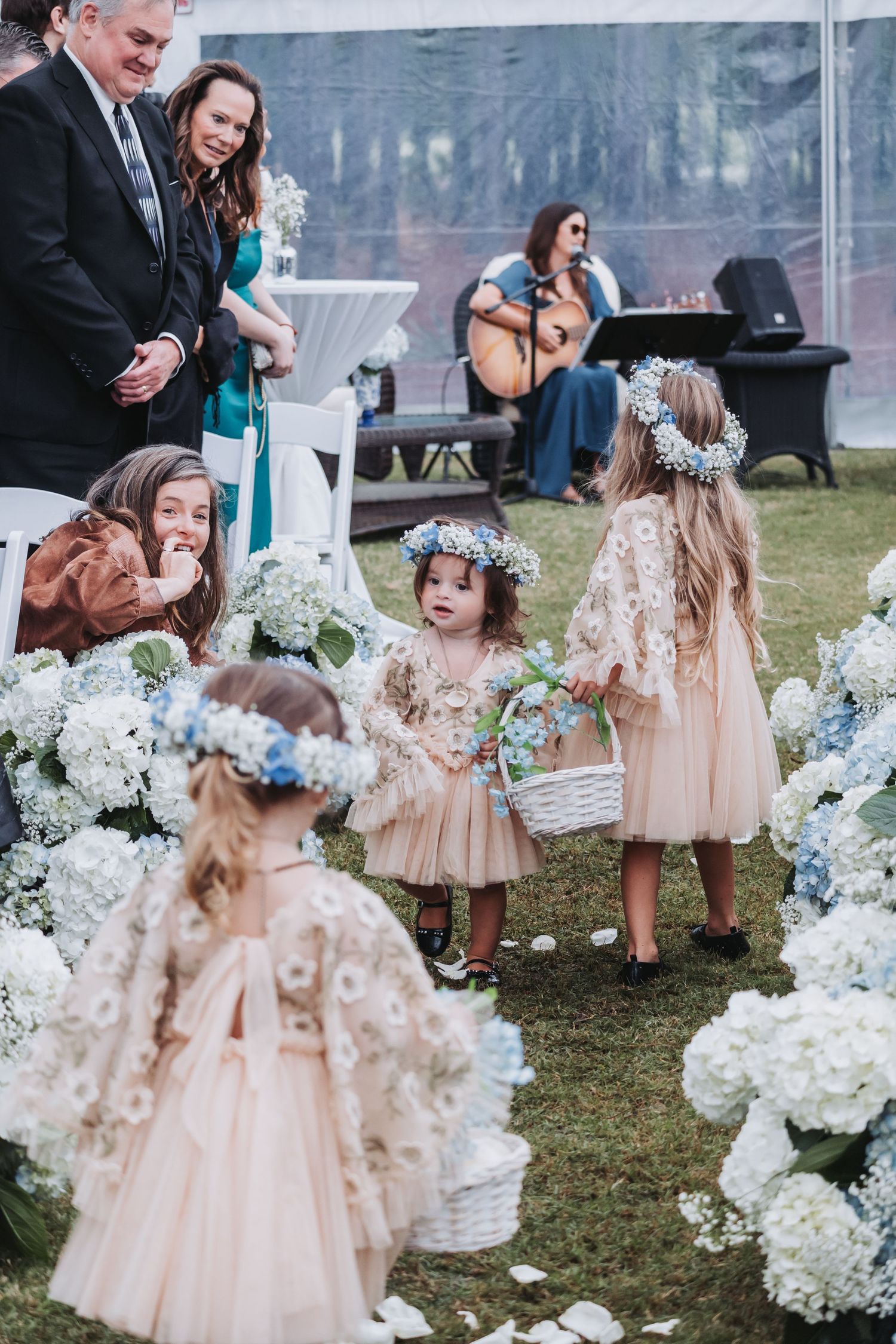 Adorable flower girls in blush dresses with floral crowns scatter petals along a hydrangea-lined wedding aisle.
