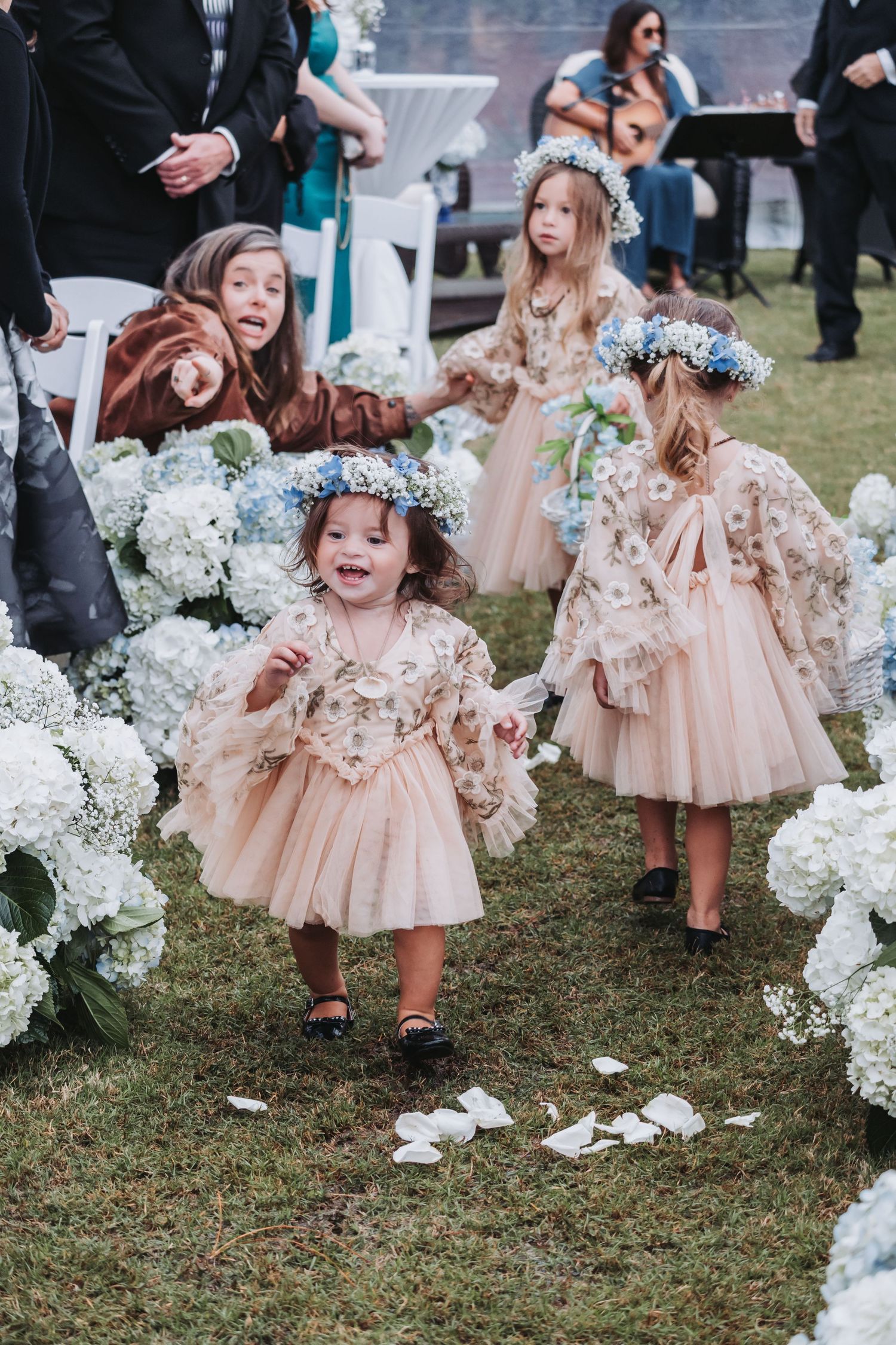 Adorable flower girls in matching blush dresses and blue floral crowns walk down an outdoor wedding aisle.