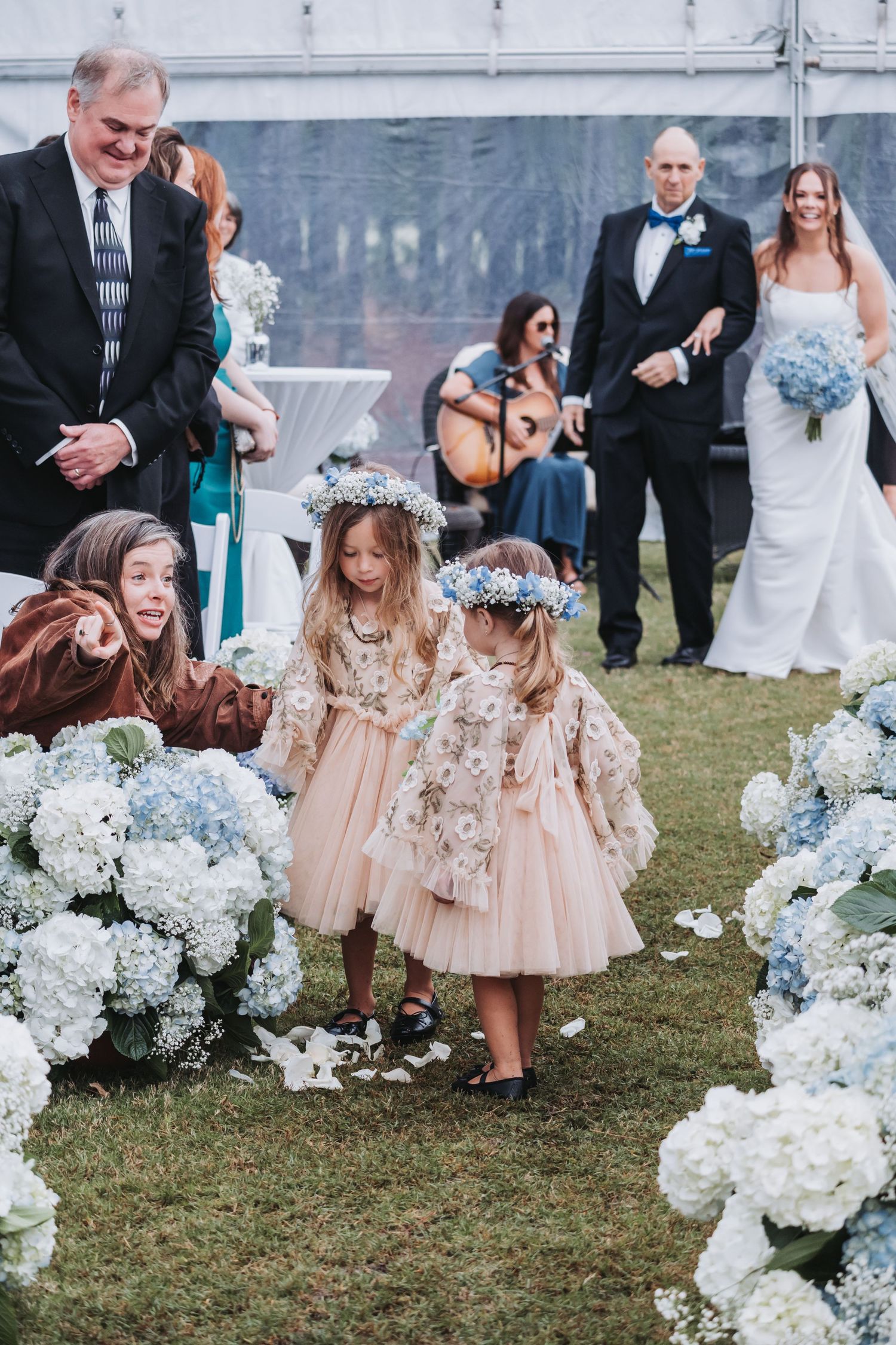 Flower girls in blush tulle dresses and floral crowns scatter petals down a hydrangea-lined wedding aisle.