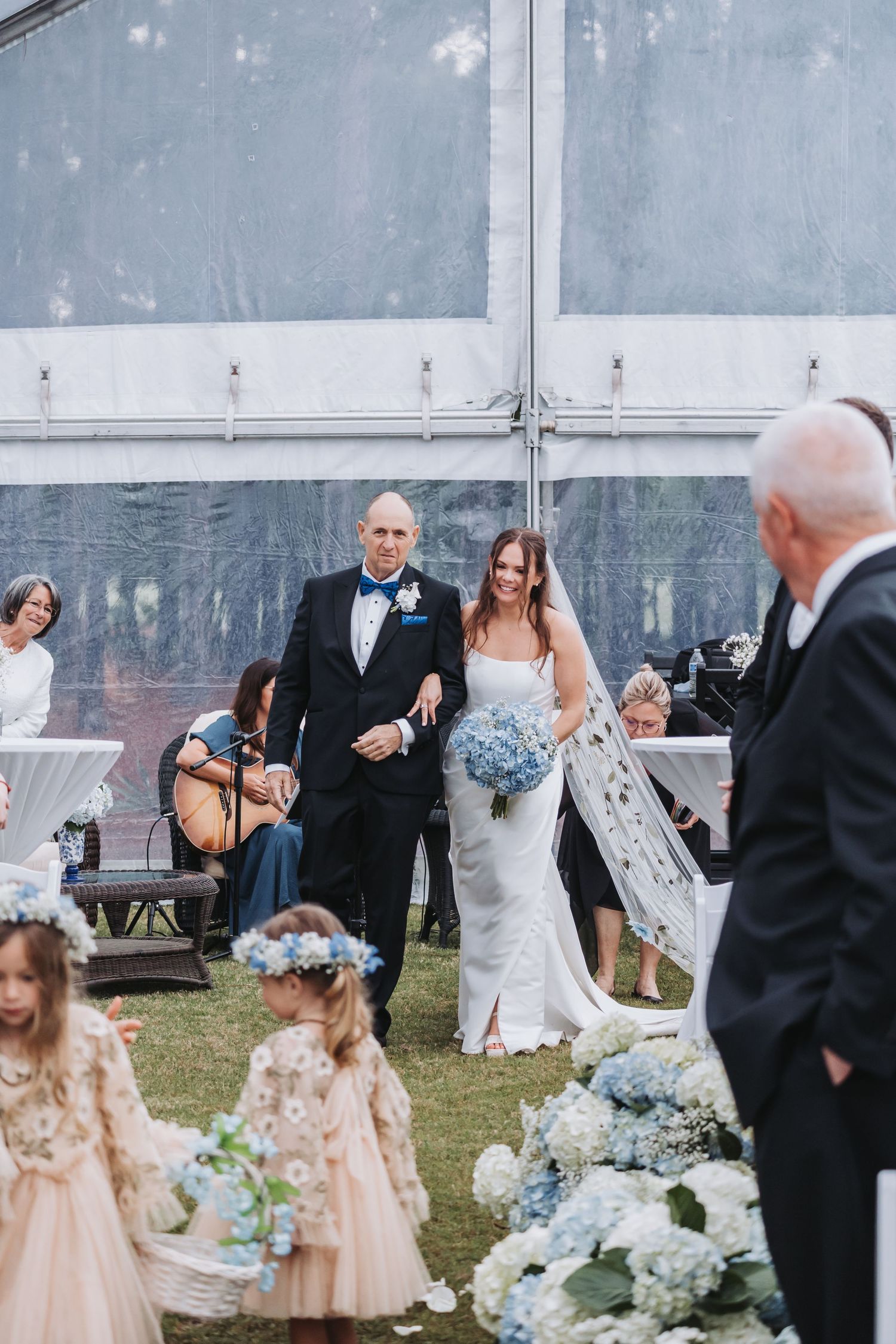 Bride and escort walking down floral-lined aisle with flower girls at outdoor tented wedding.
