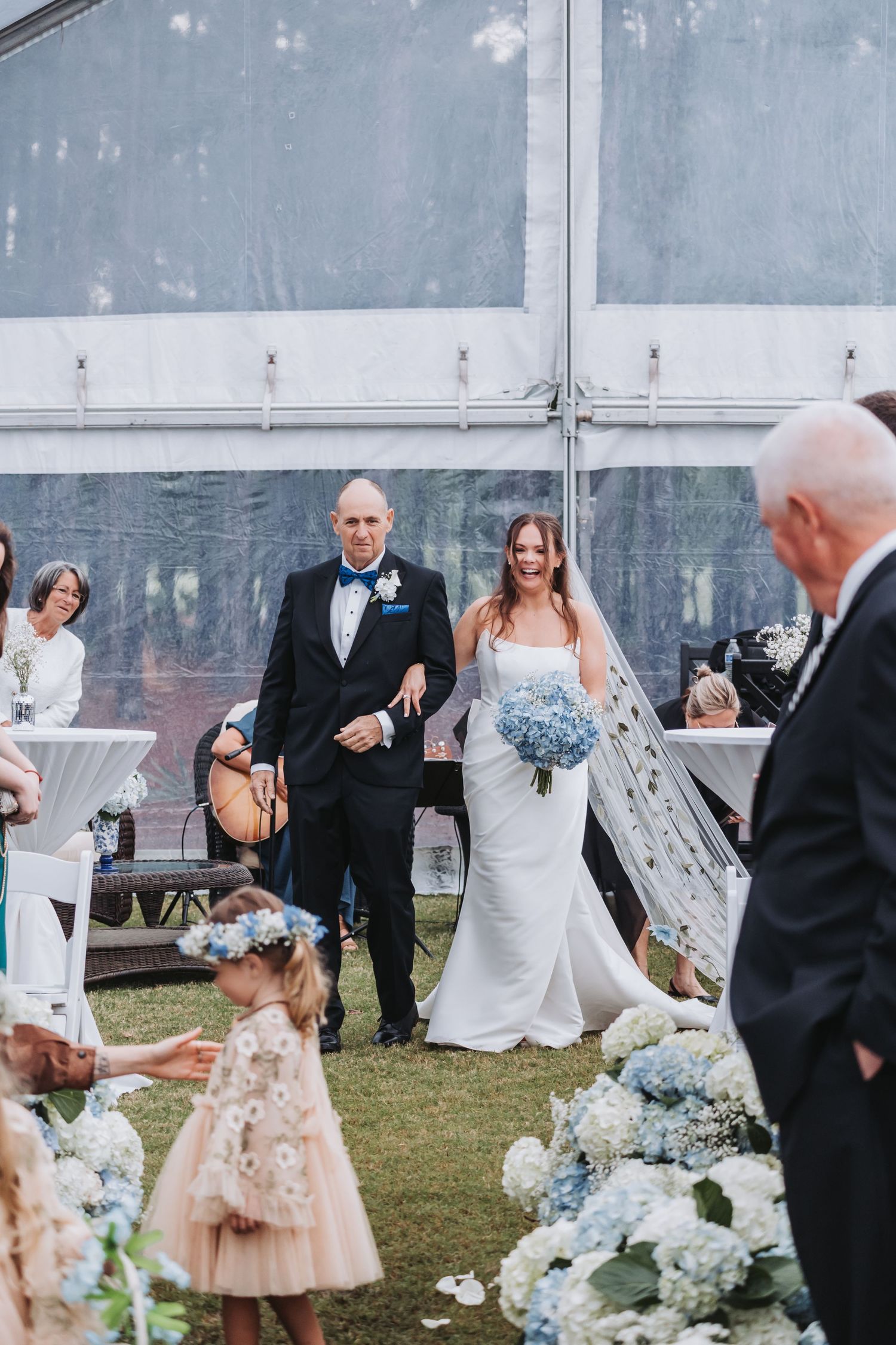 Father in tuxedo walks bride in white gown down outdoor wedding aisle lined with blue hydrangeas.