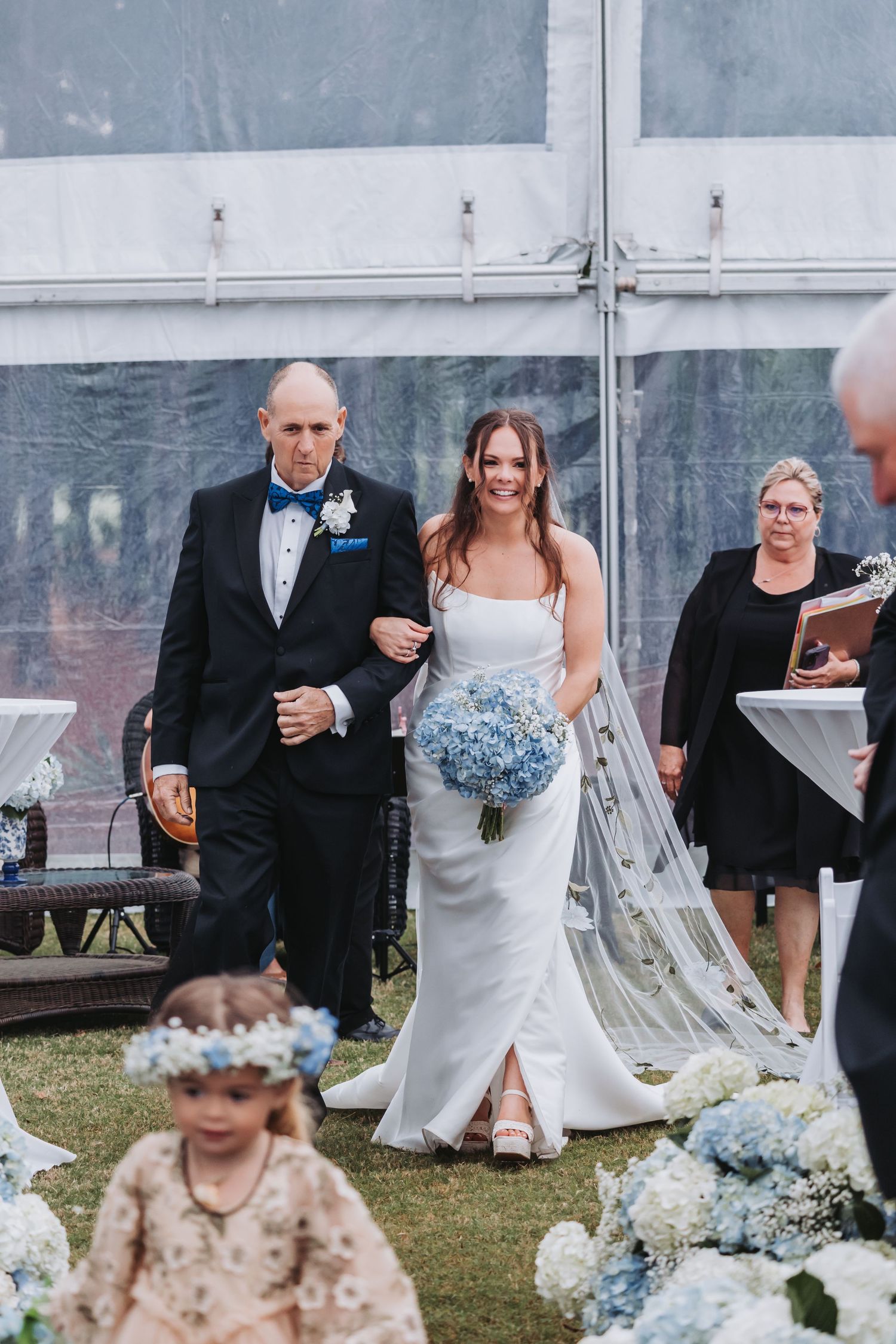 Bride walks down the aisle in a white gown holding blue hydrangeas, escorted by a man in a tuxedo.