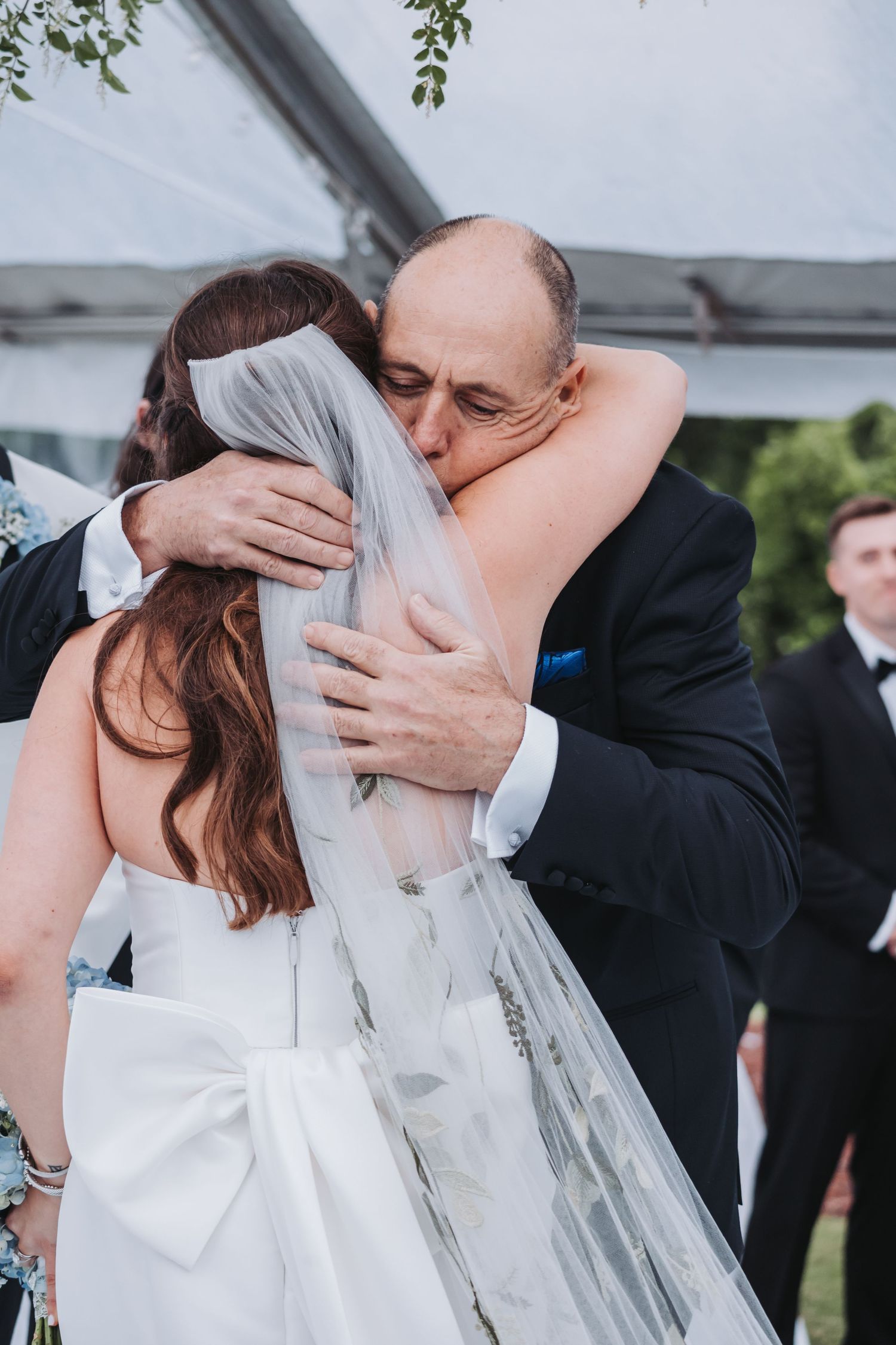 Emotional father of the bride shares tender hug with daughter in white wedding gown and veil.