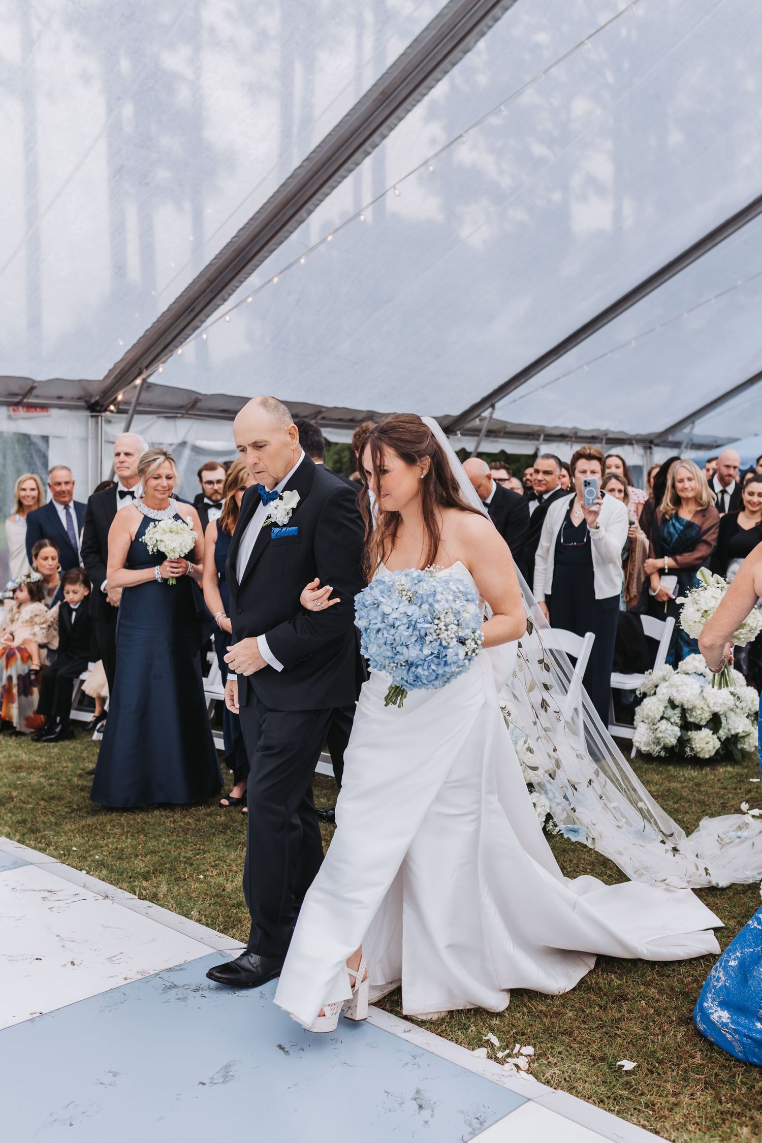 Newlywed couple walks back down aisle after ceremony under clear tent with guests applauding behind them.