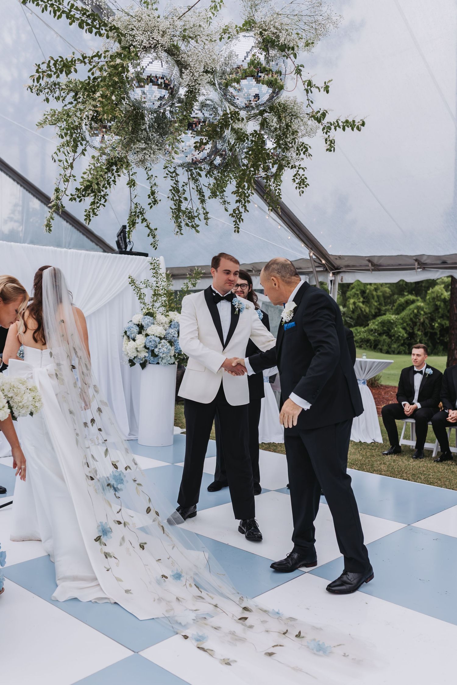 Groom in white tuxedo shakes hands with father of bride during outdoor wedding ceremony under floral chandelier.