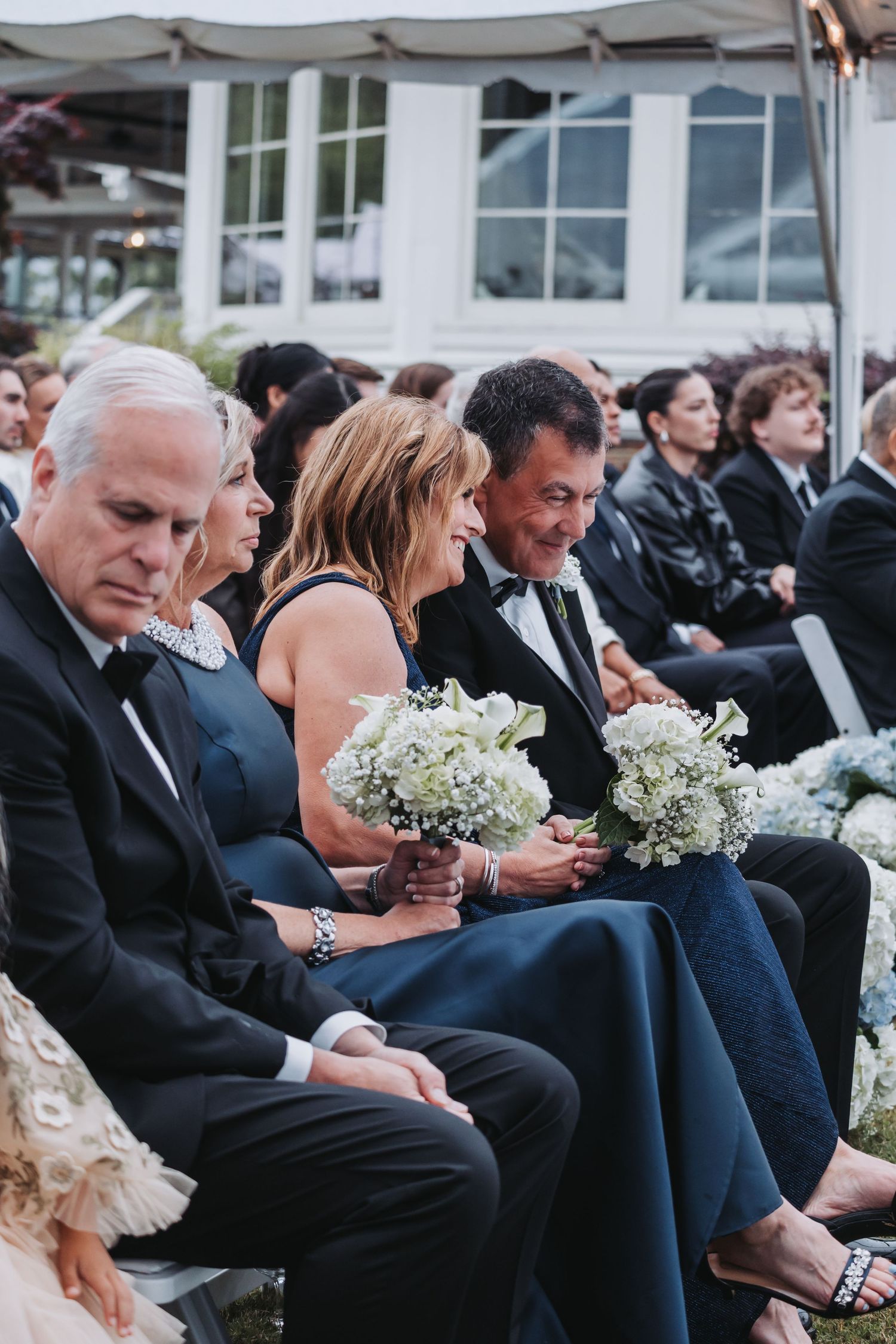 Wedding guests seated in white chairs hold white floral bouquets during outdoor ceremony under clear tent.