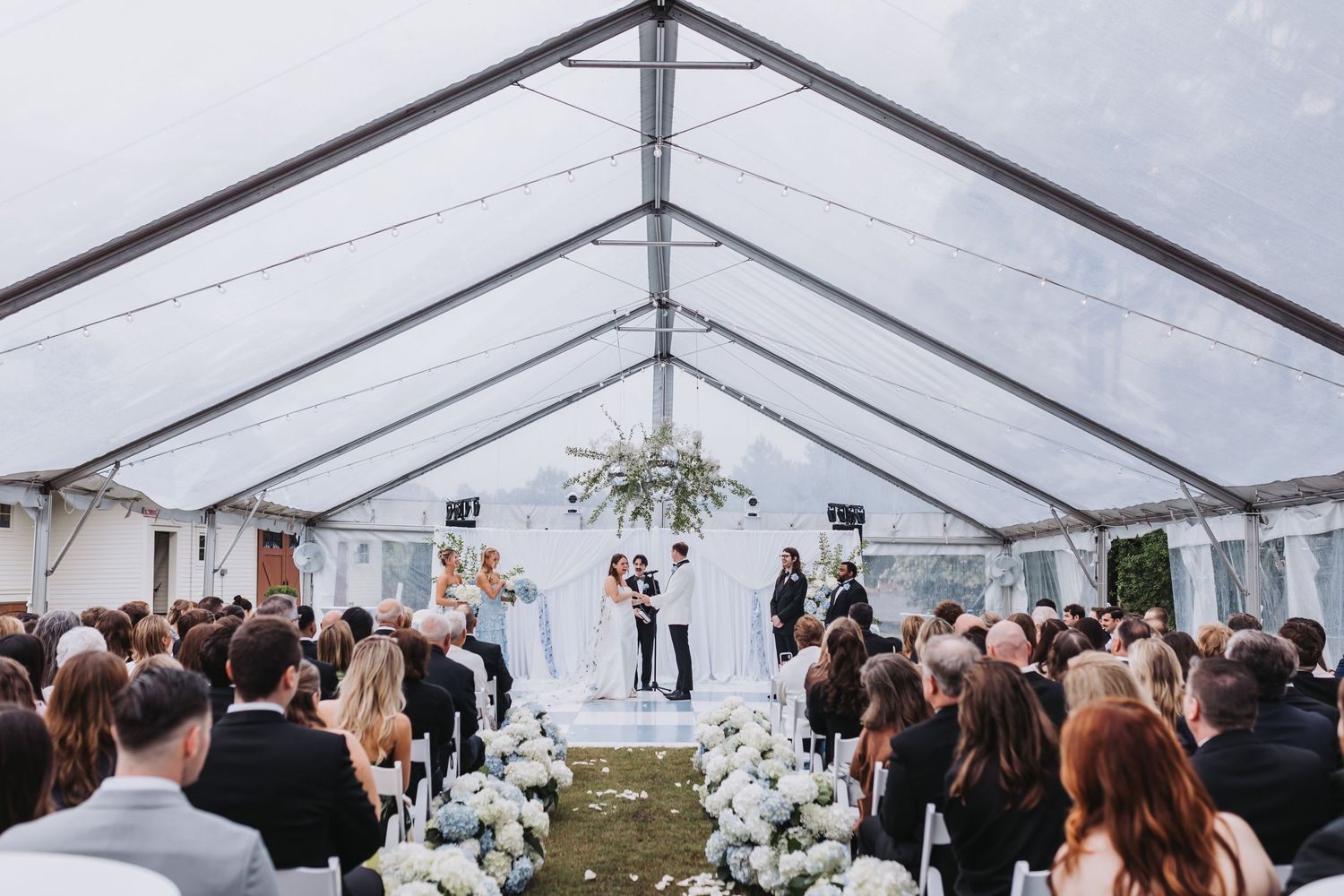 Wide shot of elegant outdoor wedding ceremony under a clear tent with white floral arrangements and large guest attendance.