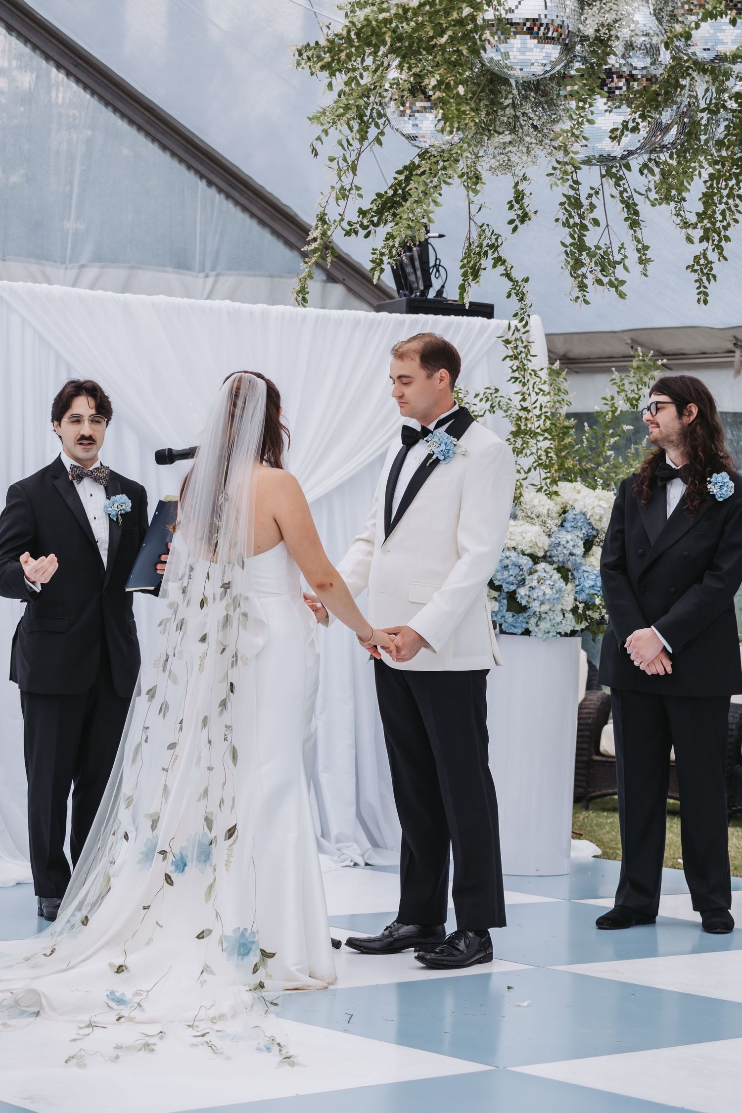 Bride in floral veil holds hands with groom in white tuxedo jacket at outdoor wedding ceremony with white drape backdrop.