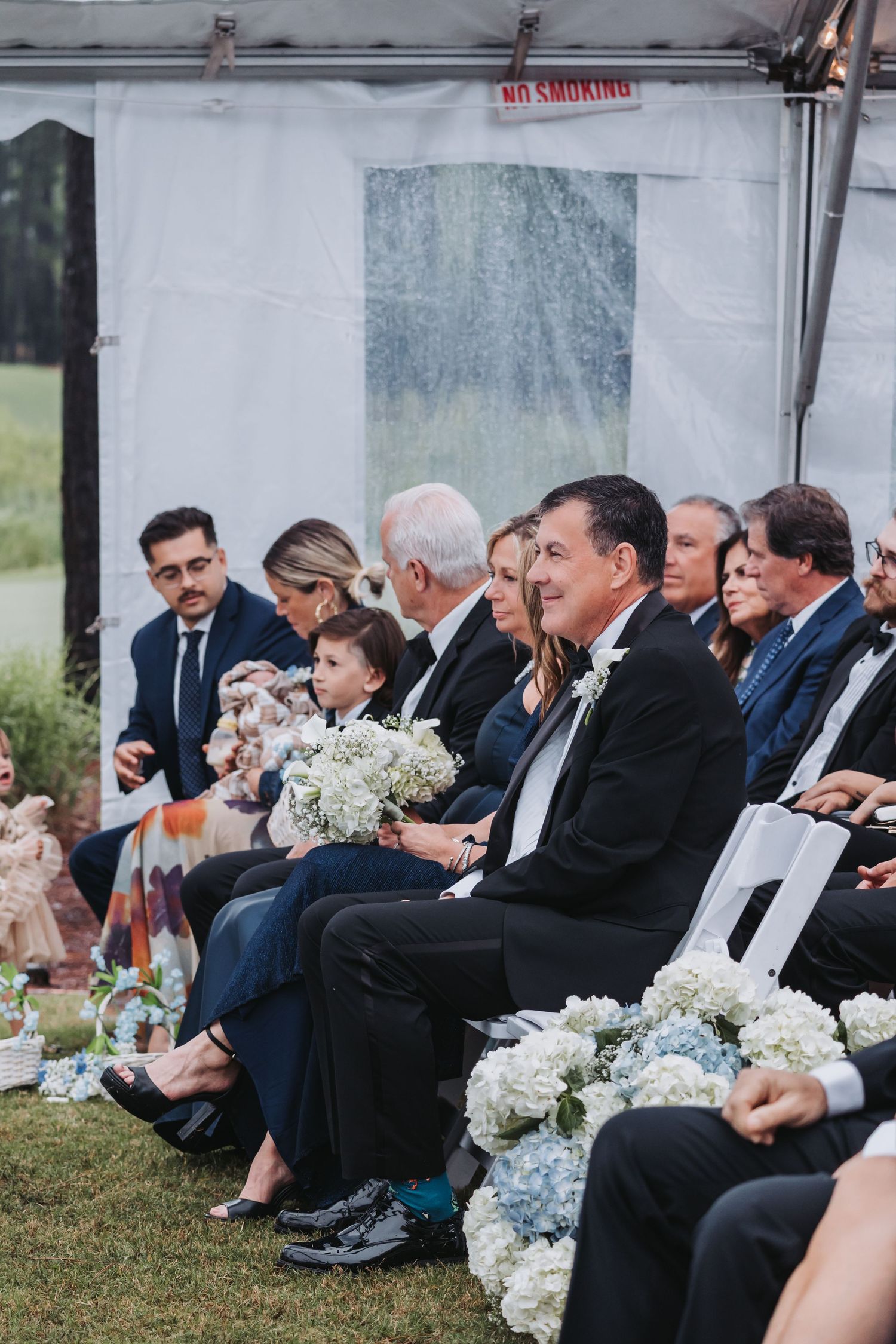 Wedding guests seated in white chairs under tent watching ceremony with blue hydrangea floral arrangements.