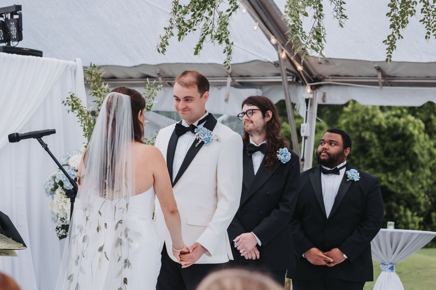 Groom in white tuxedo holds brides hands at altar during outdoor wedding ceremony under a clear tent.