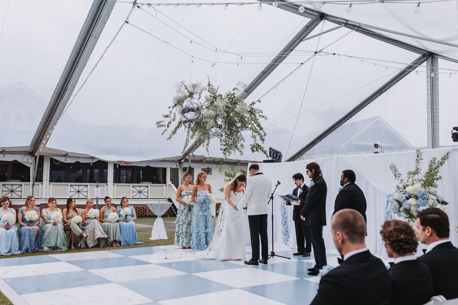 Wedding ceremony under clear tent with bride and groom at altar, bridesmaids in blue dresses seated nearby.