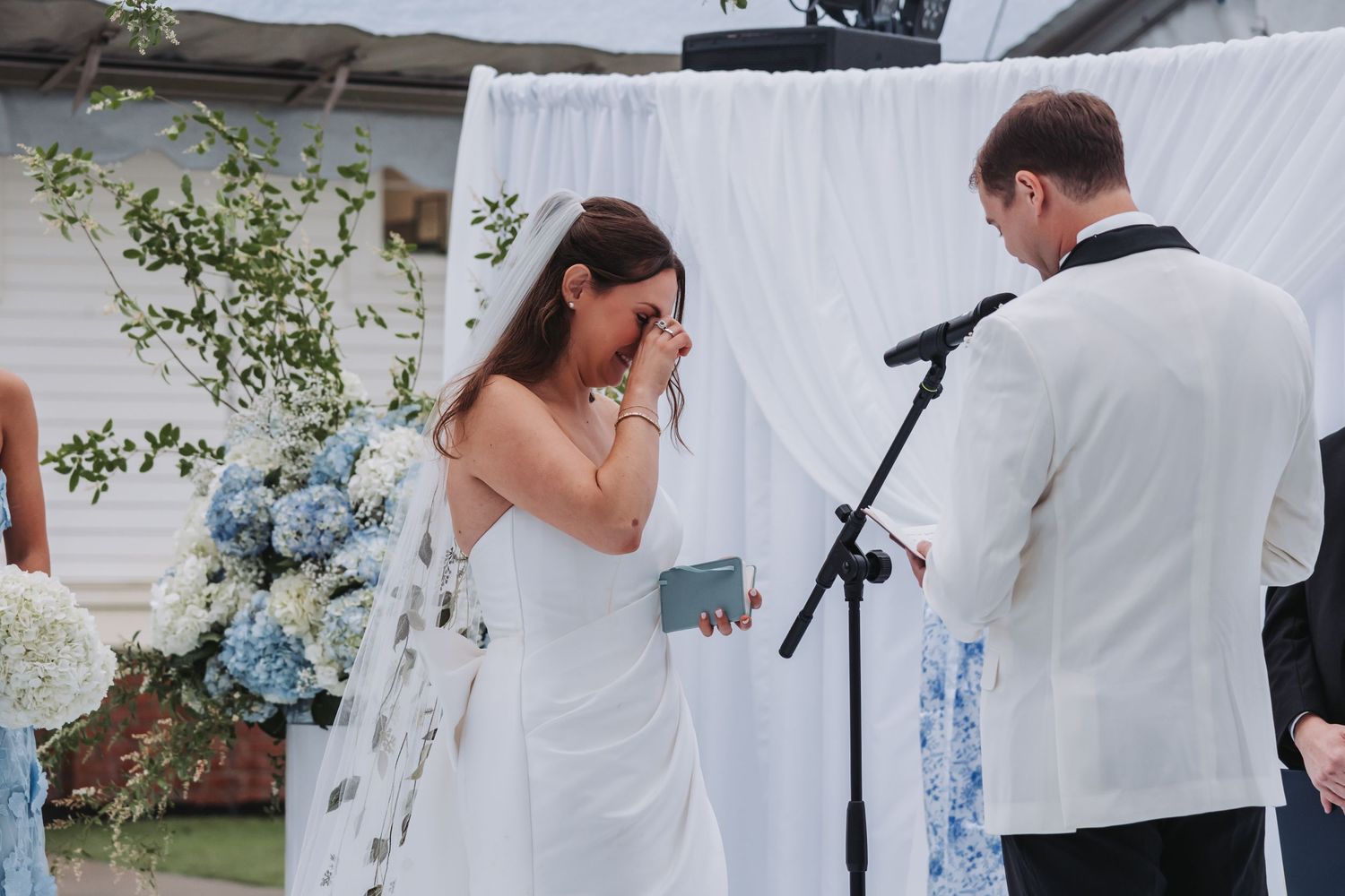 Emotional bride wipes tears while groom reads wedding vows at outdoor ceremony decorated with blue hydrangeas.