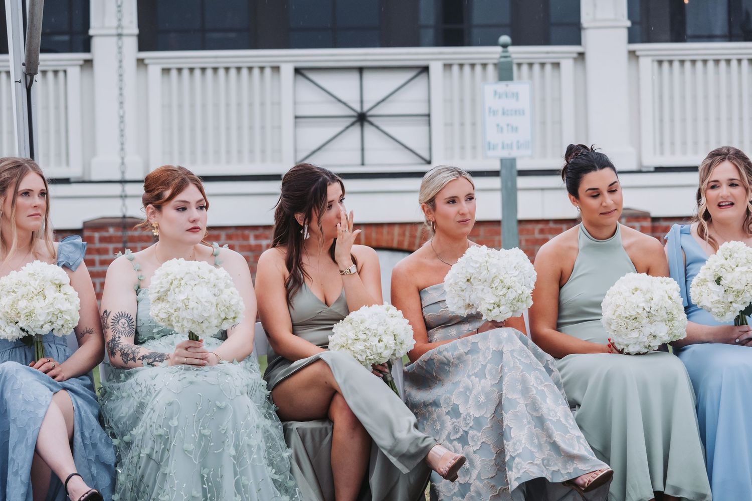 Bridesmaids in mismatched blue and sage dresses holding white hydrangea bouquets seated during outdoor wedding ceremony.