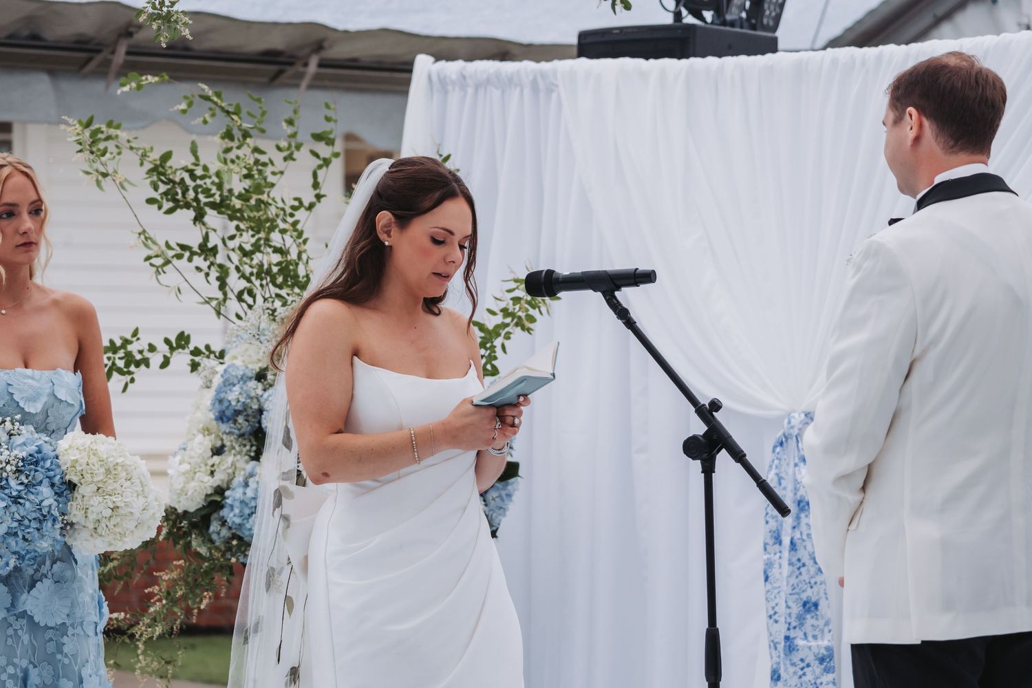 Emotional bride in strapless white gown reads heartfelt vows at outdoor wedding altar with white drape backdrop.