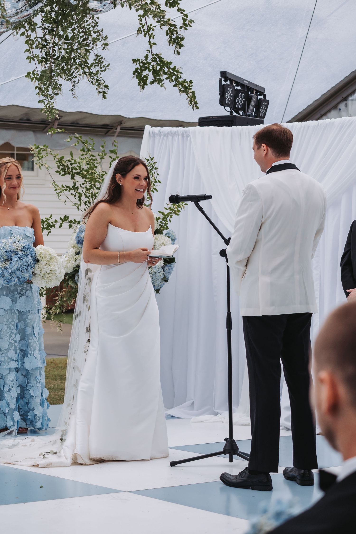 Bride smiling at groom during outdoor wedding ceremony with white drape backdrop and blue floral arrangements.