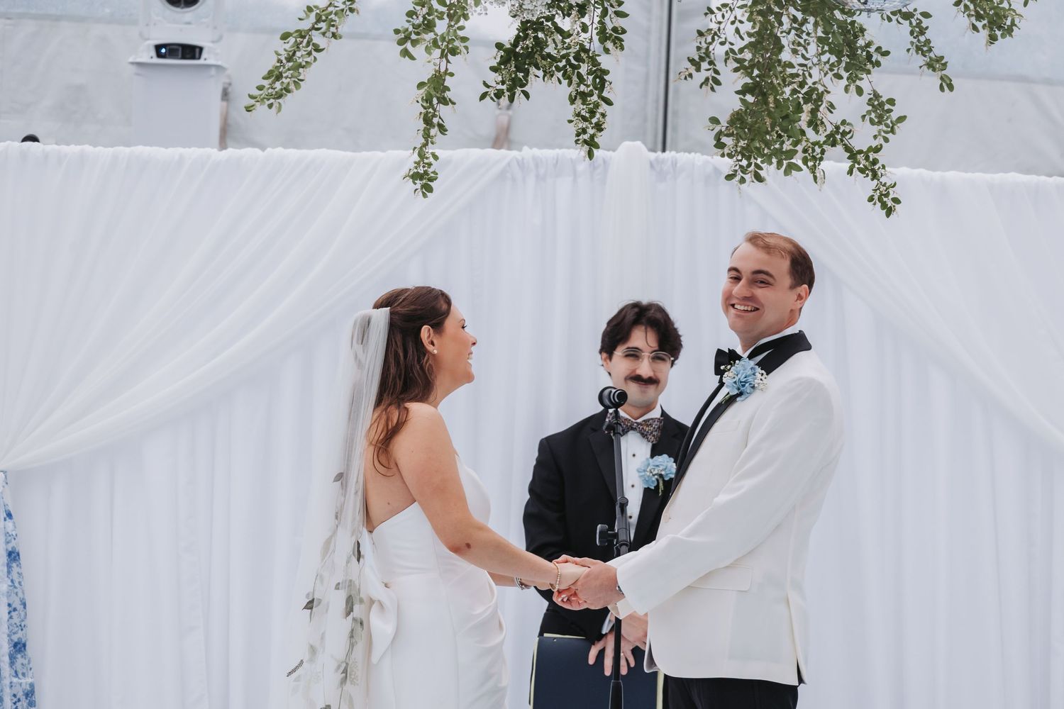 Bride and groom hold hands at outdoor wedding ceremony with officiant, white drape backdrop, and greenery.