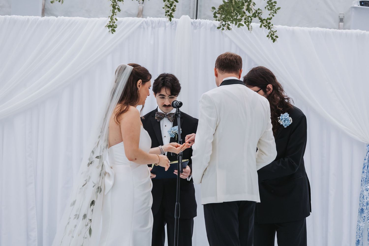 Bride and groom exchange rings with officiant and wedding party at elegant outdoor ceremony with white drape backdrop.