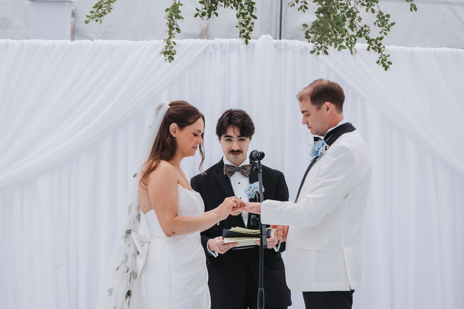 Bride and groom exchange rings during outdoor wedding ceremony with officiant at white draped altar.