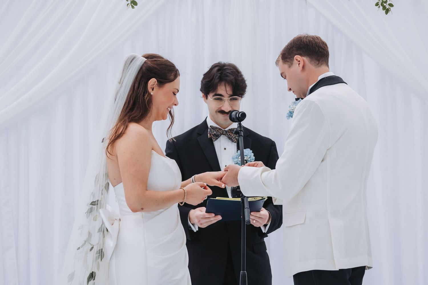 Bride and groom exchange rings during outdoor wedding ceremony with officiant holding microphone.