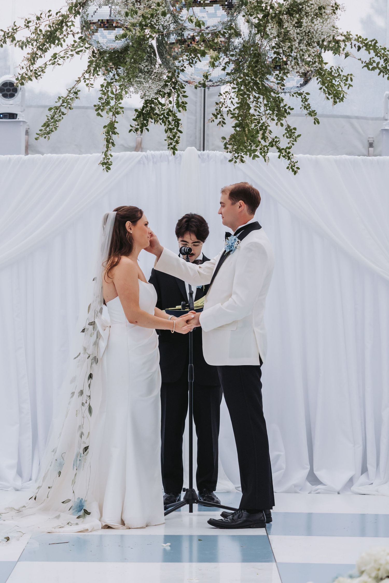 Bride and groom exchange vows at elegant outdoor wedding ceremony with white draping and greenery arch.