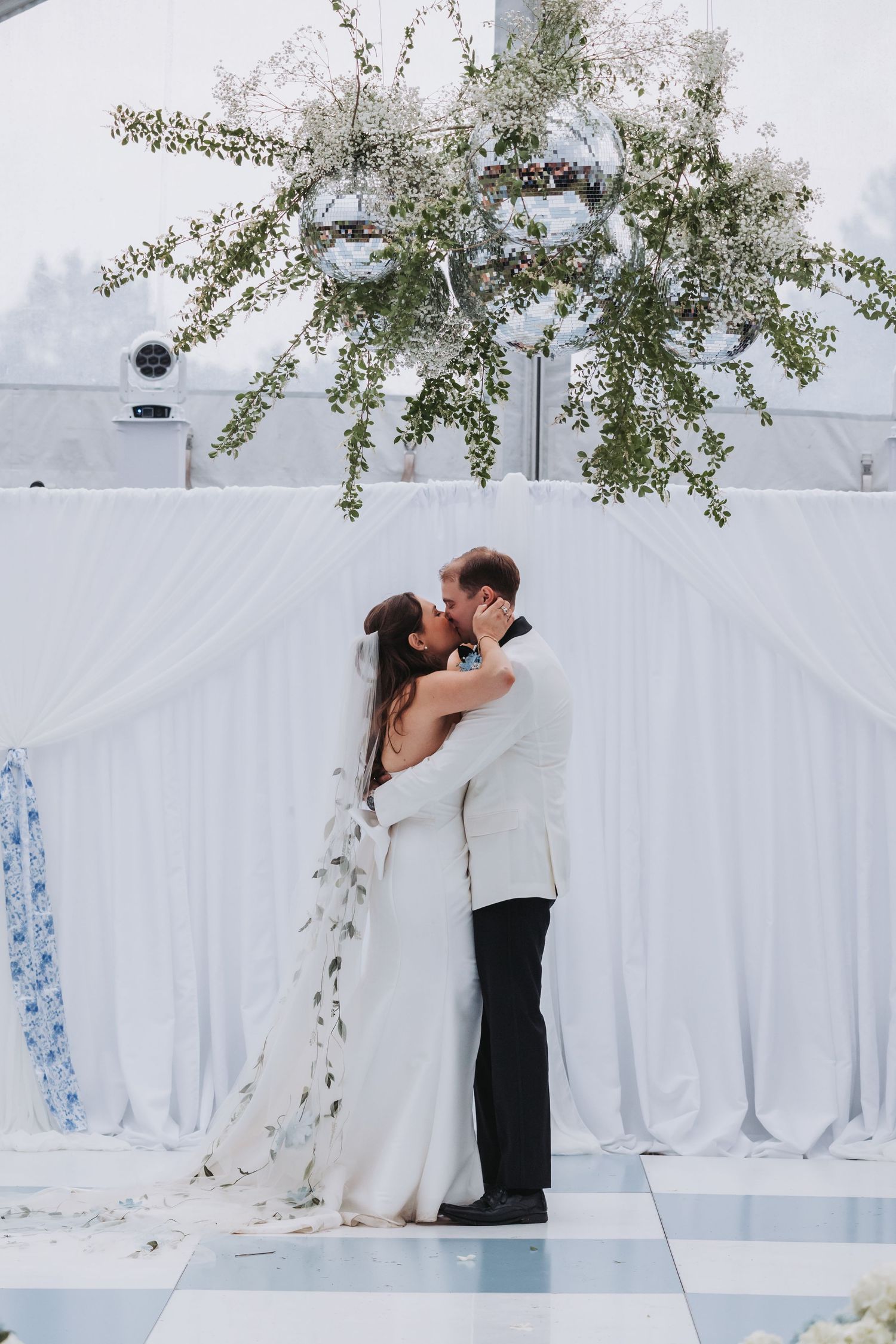 Bride and groom share first kiss at elegant white draped altar with greenery and disco ball overhead.