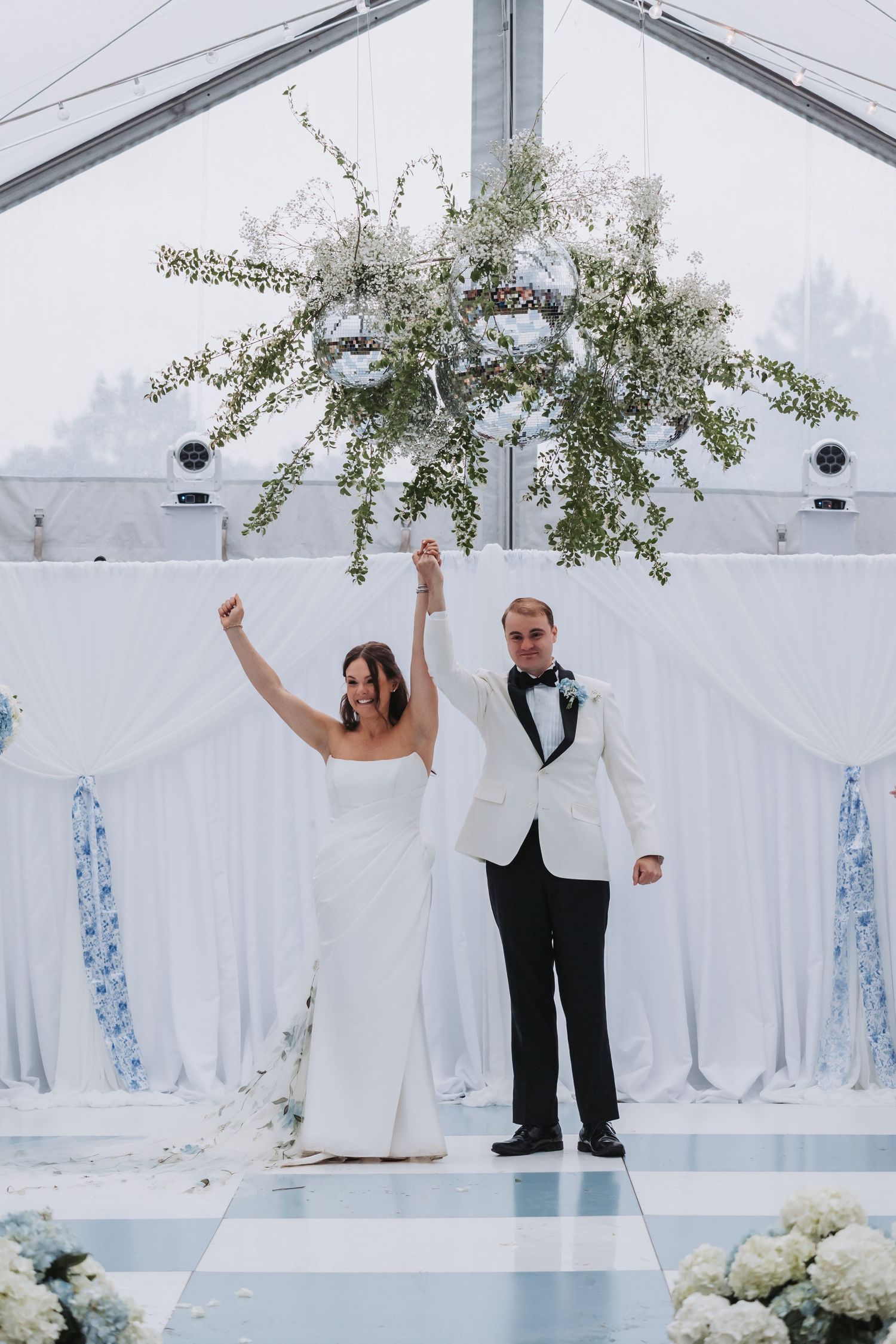 Bride and groom celebrate just married moment raising hands under floral chandelier in elegant white tented wedding venue.