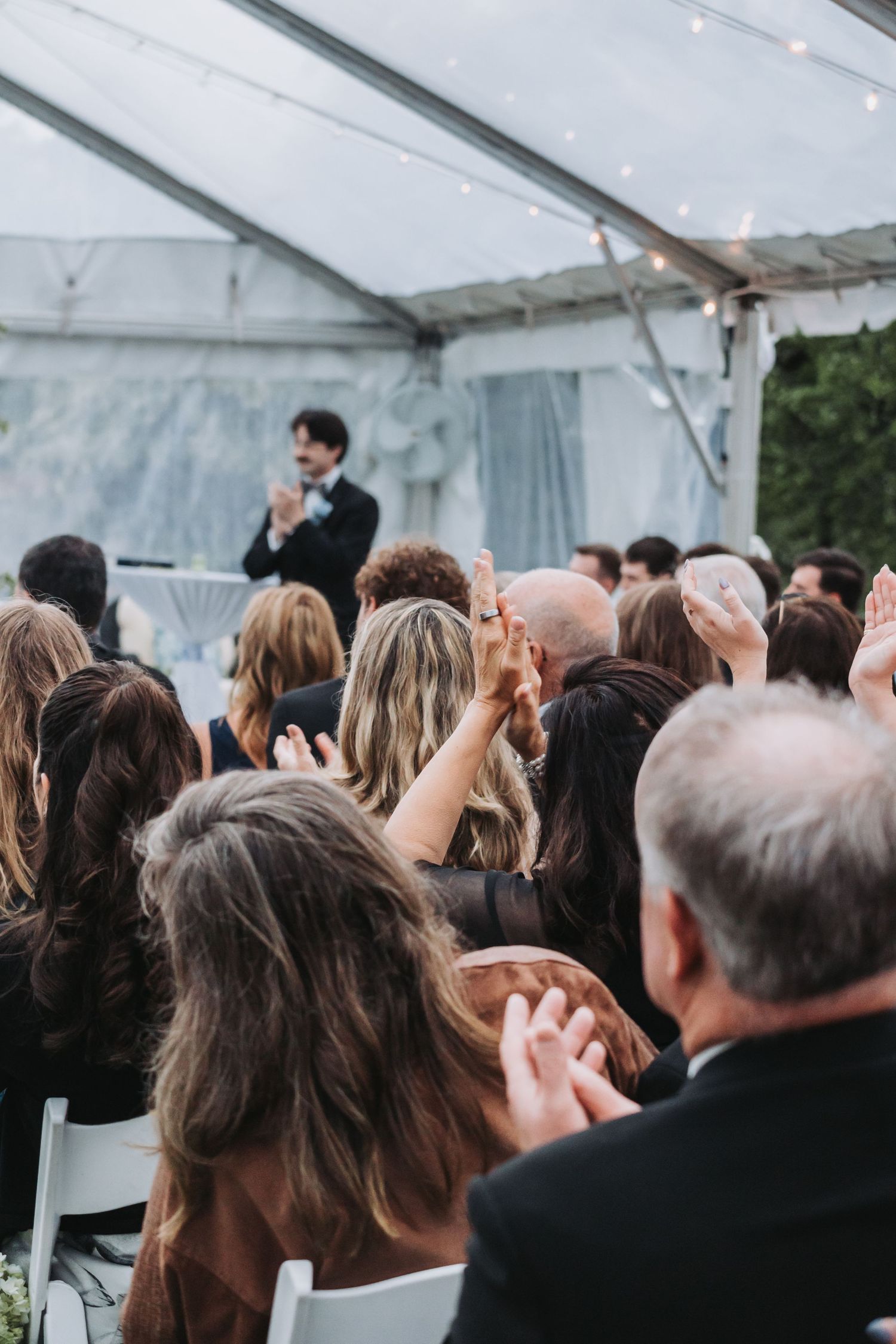 Guests clapping and raising hands at an outdoor wedding reception under a white tent with string lights.