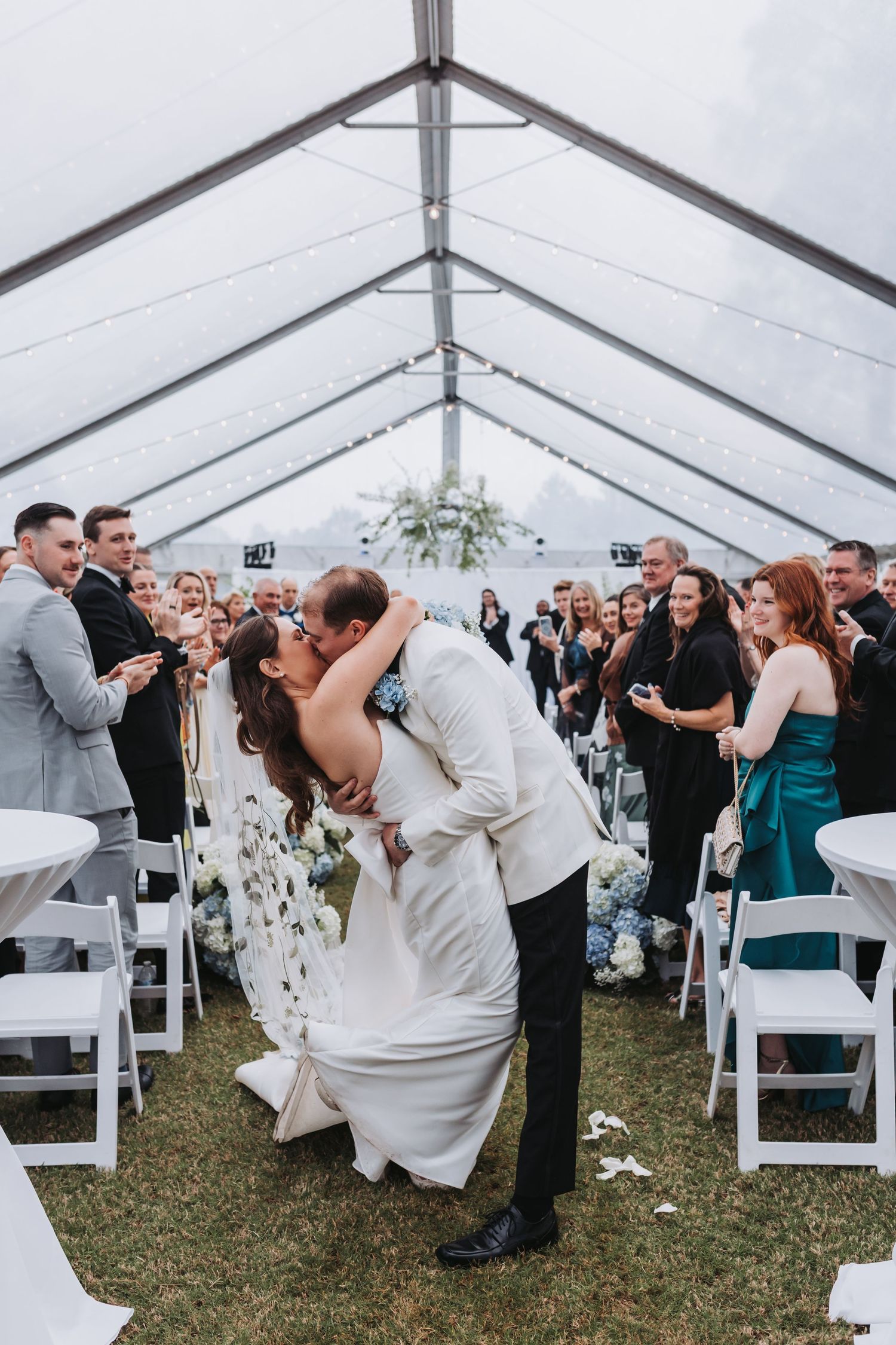 Groom dips bride for a passionate kiss at the end of the wedding aisle surrounded by cheering guests.