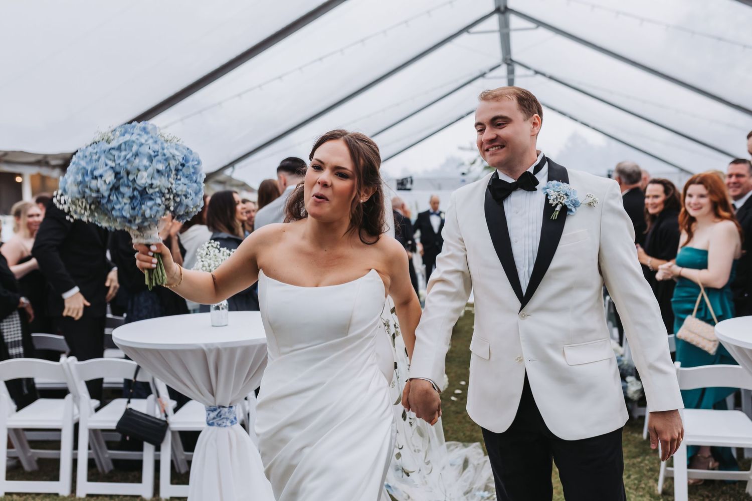 Bride in strapless gown and groom in white tuxedo walk joyfully down aisle under clear tent at outdoor wedding.