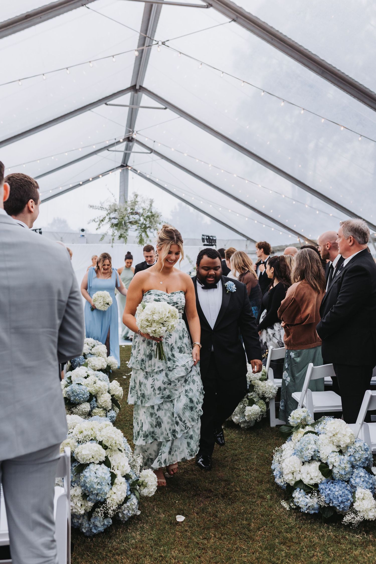 Bridesmaid in floral dress walks down aisle lined with blue and white hydrangeas under clear tent.