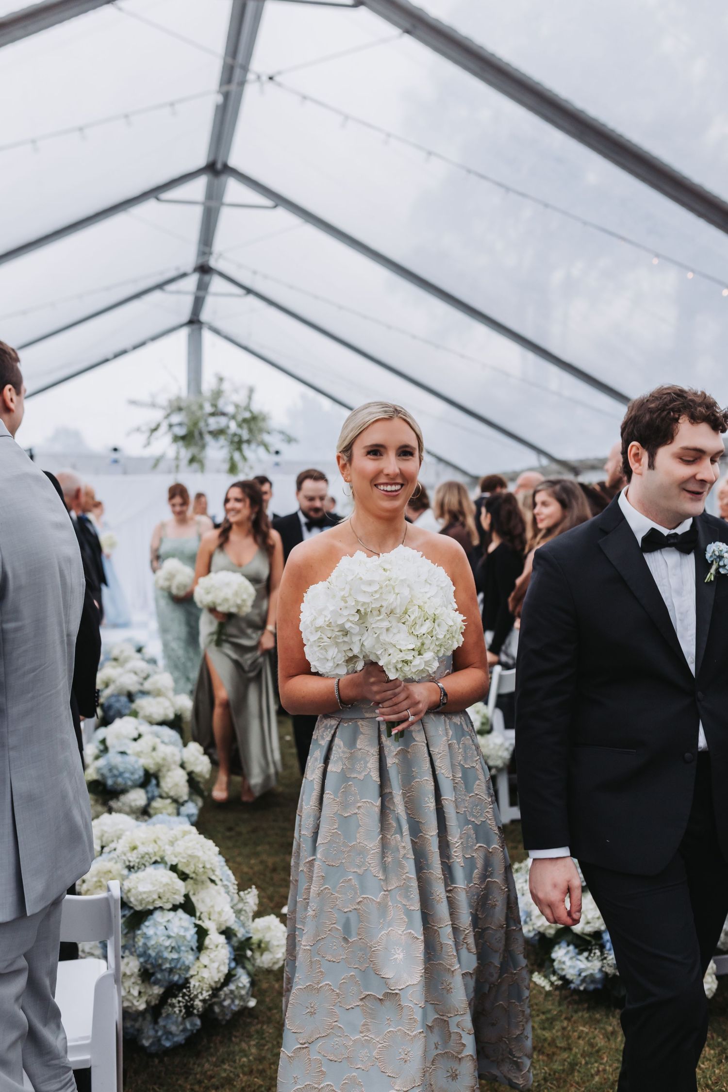Bridesmaid in silver brocade gown holds white hydrangea bouquet while walking down aisle in elegant tent wedding ceremony.