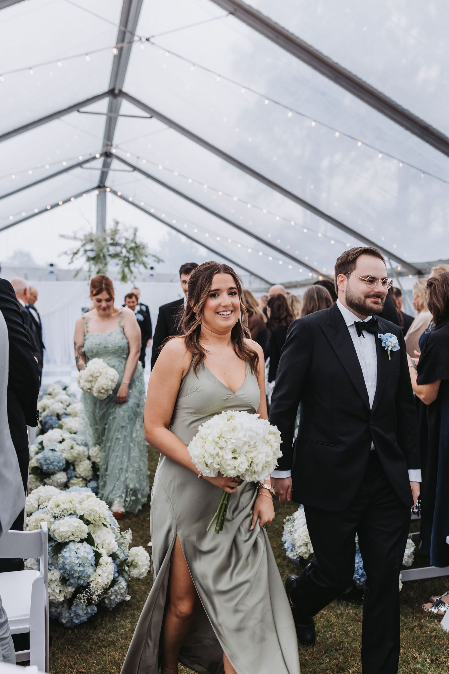 Bridesmaid in sage green satin dress walks down wedding aisle holding white hydrangea bouquet under a clear tent.