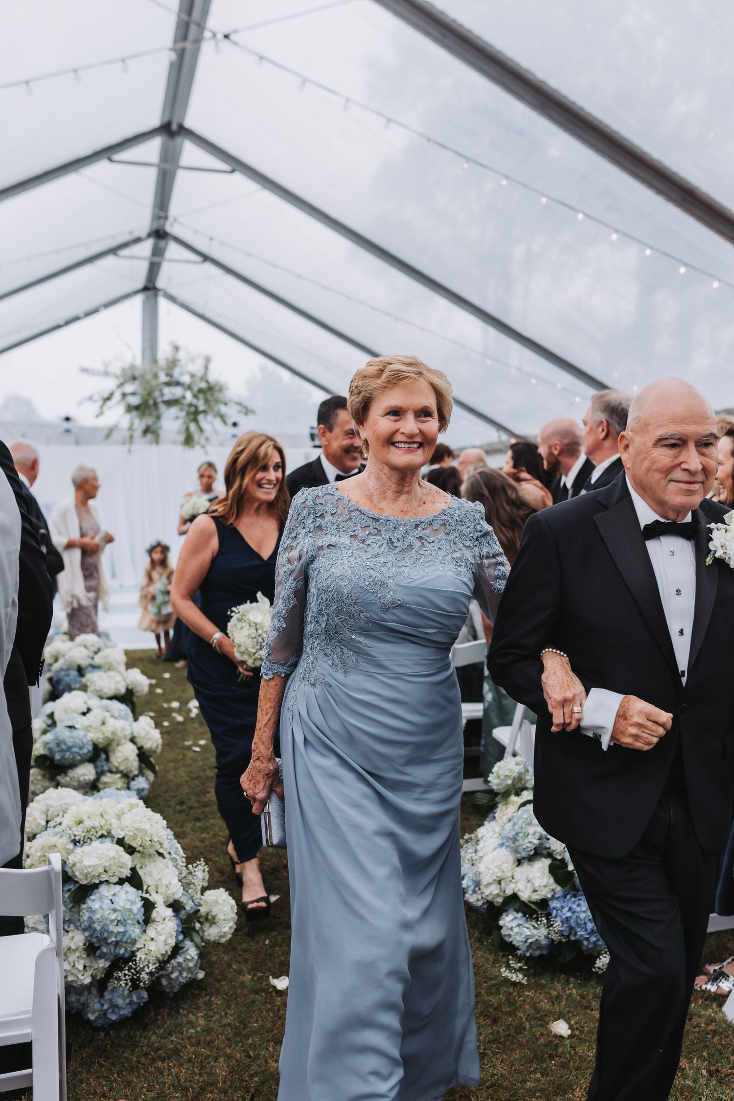 Elegant mother of the bride in blue lace gown walks down flower-lined wedding ceremony aisle.