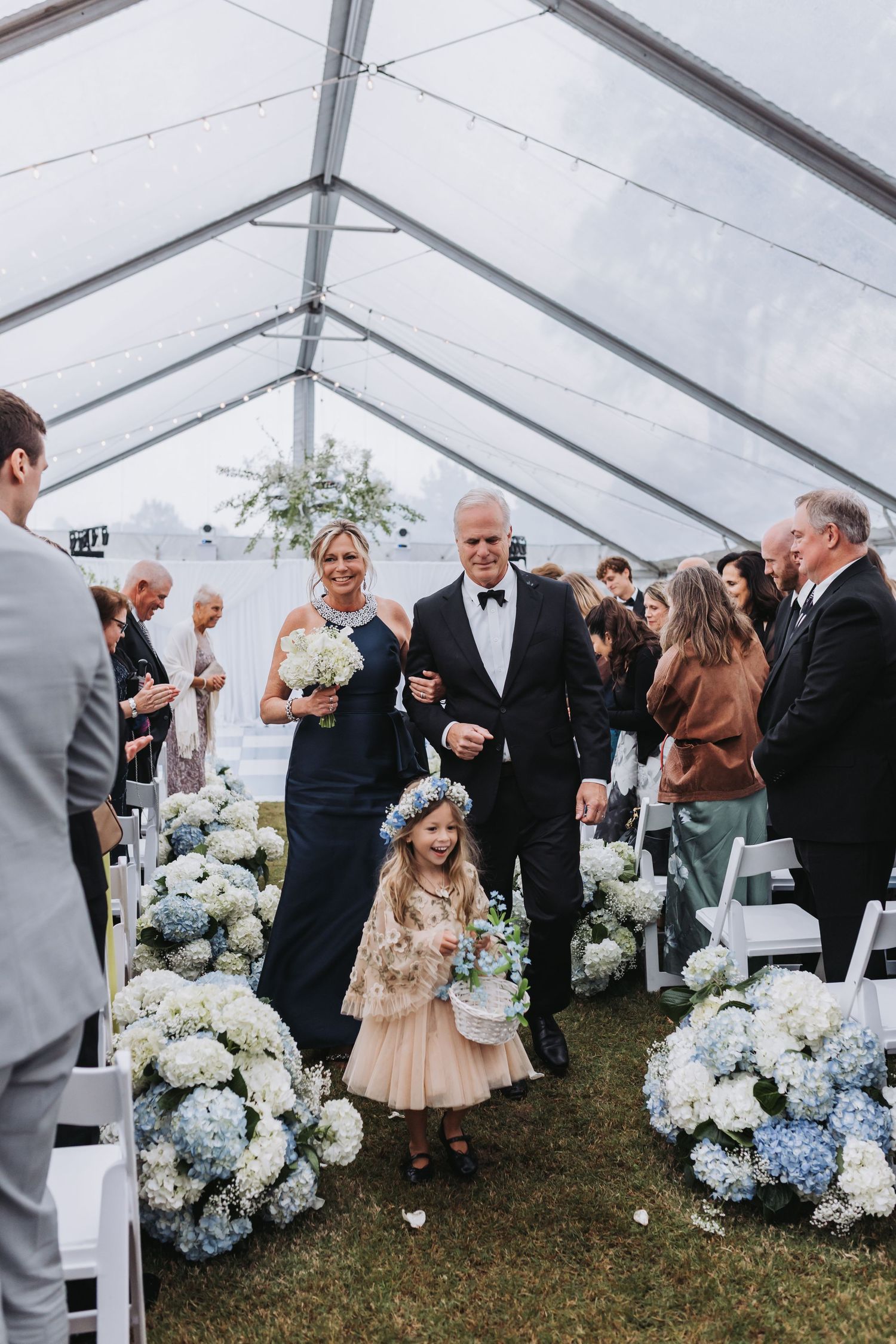 Adorable flower girl in floral crown walks down aisle with family past blue hydrangea arrangements.