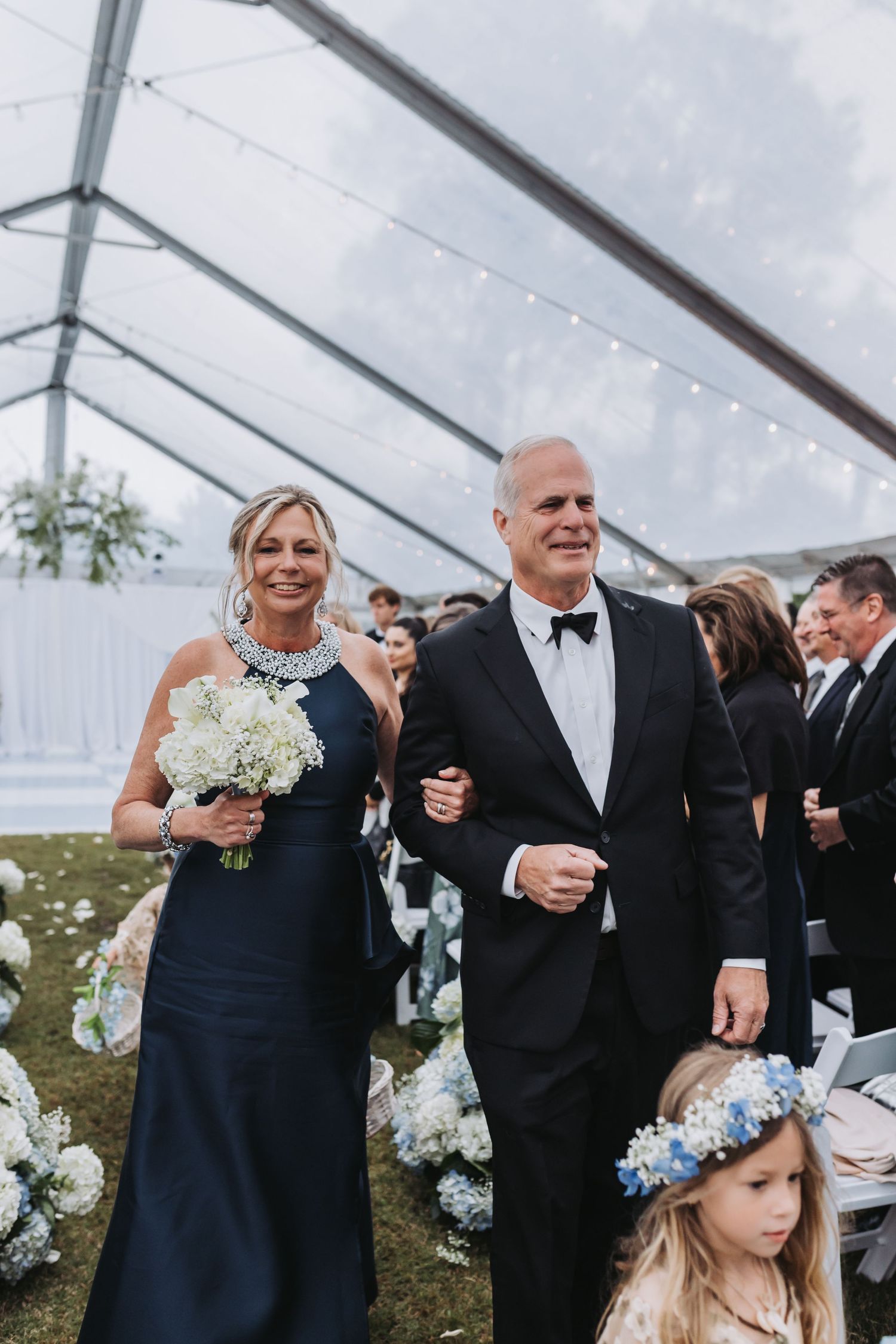 Elegant wedding processional with bridesmaid in navy gown holding white bouquet walking arm-in-arm with groomsman under tent.