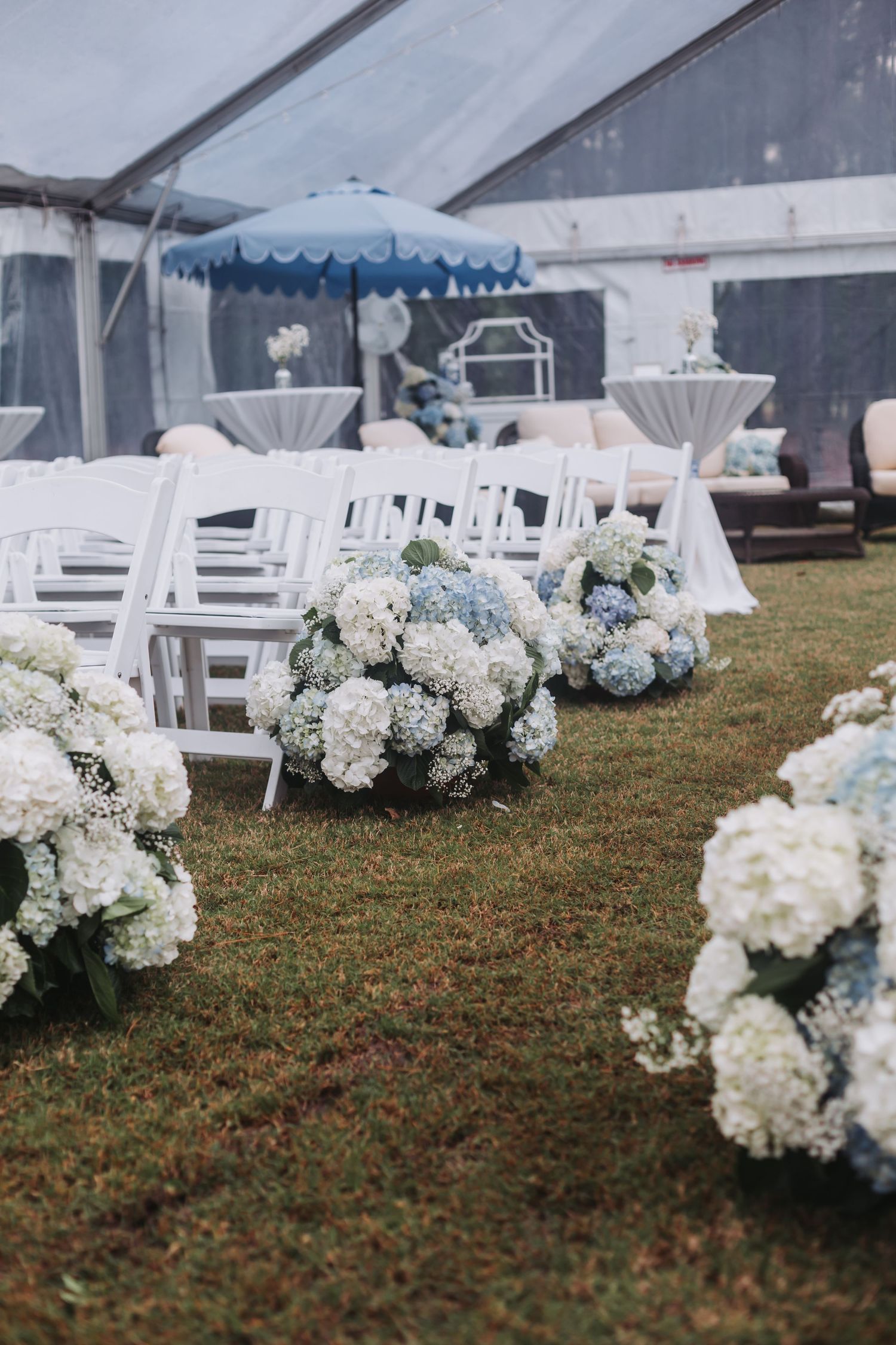 Elegant outdoor wedding ceremony aisle lined with blue and white hydrangea floral arrangements on green grass under a white tent.
