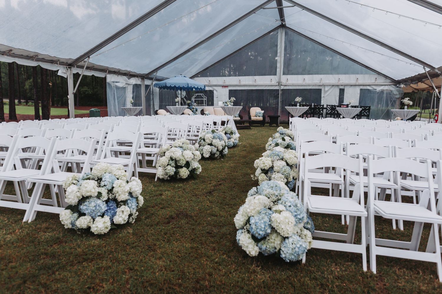 White folding chairs line a grass aisle adorned with blue and white hydrangea floral arrangements under a clear wedding tent.
