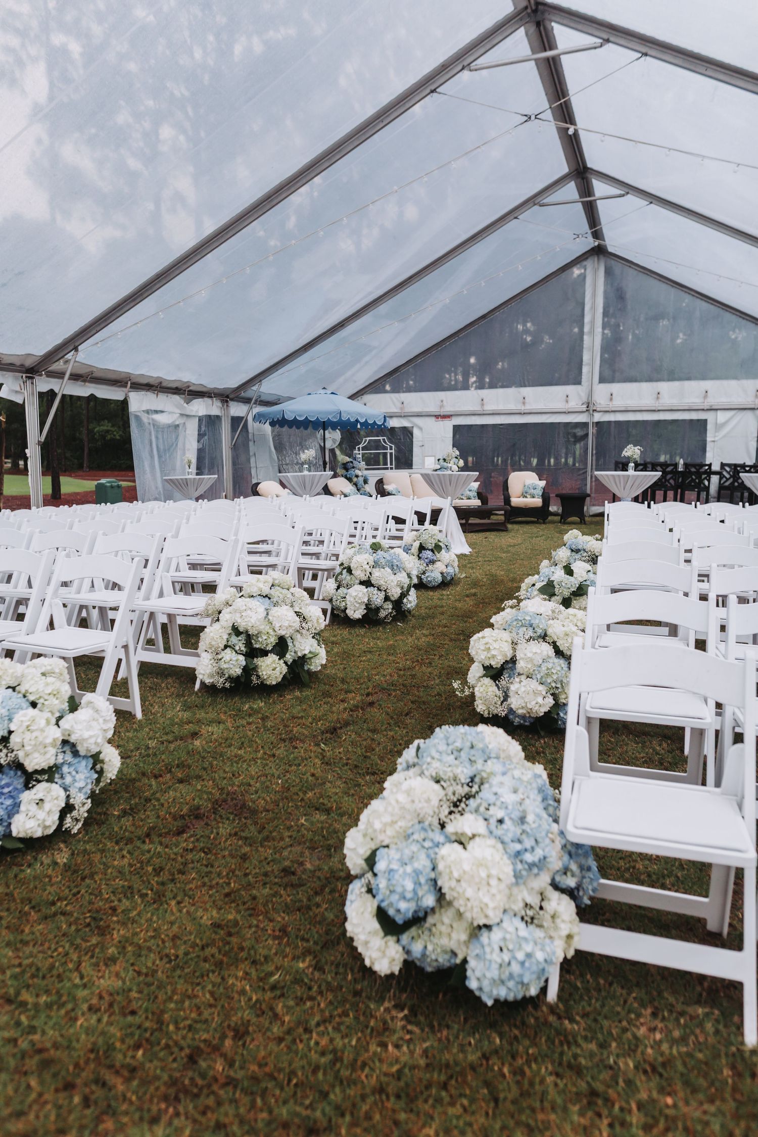 White folding chairs line a grass aisle adorned with blue and white hydrangea bouquets under a clear wedding tent.