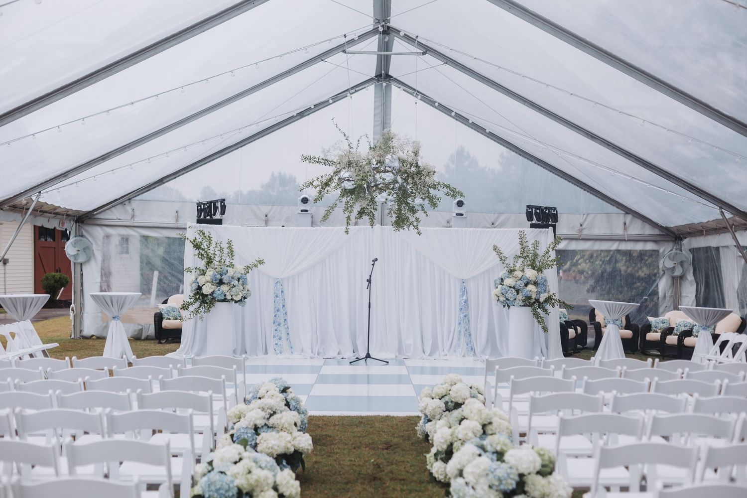 Elegant white wedding tent ceremony setup with hydrangea aisle flowers and floral arch backdrop.