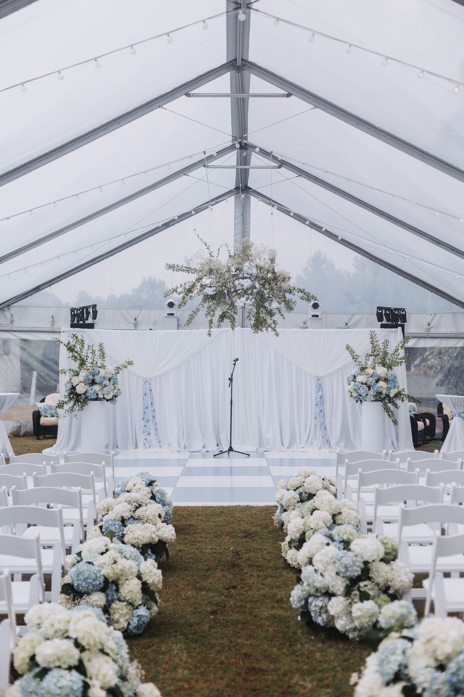 White wedding ceremony tent with hydrangea-lined aisle, draped altar, and string lights overhead.