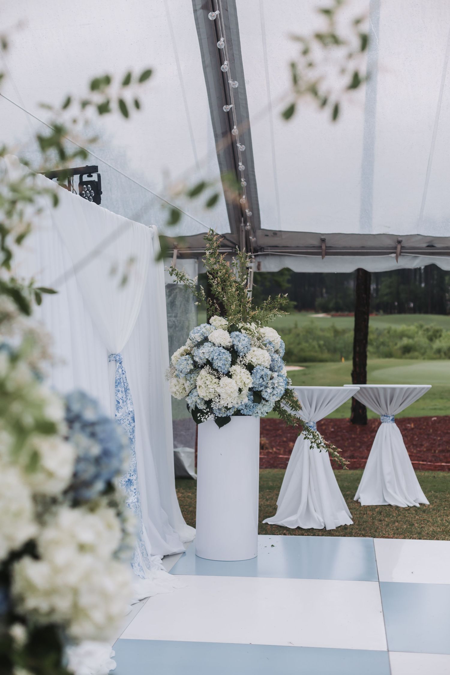 White cylinder vase with blue and white hydrangea arrangement on checkered wedding ceremony floor.
